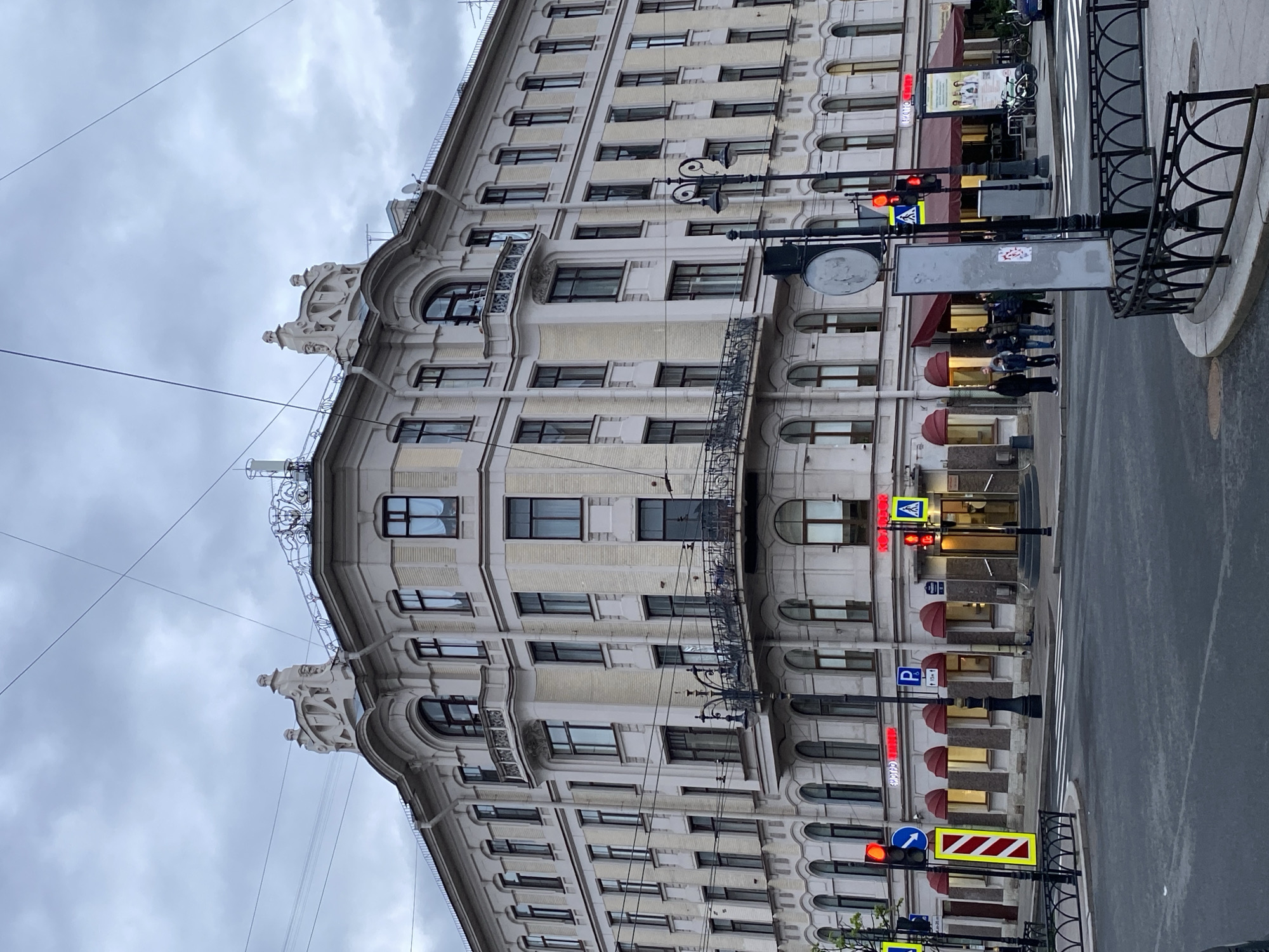 The image shows a multi-story building in Saint Petersburg, Russia. The building is light-colored with numerous arched windows. The architecture features decorative elements on the roofline and wrought-iron balconies. A street runs along the side of the building. On the street, there are pedestrian crosswalk signs, traffic lights, and lampposts. Several people are standing on the sidewalk in front of the building. The sky is overcast.