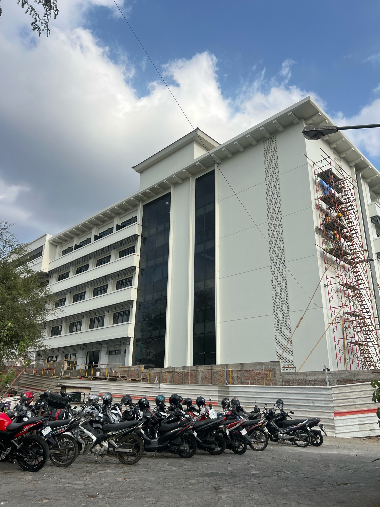 The image depicts the exterior of a white, multi-story building, likely a modern educational or administrative facility, under construction or renovation. Scaffolding is erected on the right side of the building, where several individuals in hard hats and safety vests are visible, suggesting ongoing work. The building features dark, reflective windows arranged in horizontal bands on each floor, interspersed with white panels. Decorative patterned elements are visible on the facade, particularly a vertical strip of intricate white lattice work.
In the foreground, a row of parked motorcycles, predominantly scooters, are lined up on a paved surface. These motorcycles are of various colors, including red, black, and silver. Many are equipped with helmets, indicating their use by building occupants or workers. A white metal barrier, possibly for crowd control or construction site demarcation, separates the motorcycles from the building's lower level, where exposed brickwork and construction materials are visible.
The environment is outdoors, with a clear blue sky dotted with scattered white clouds. The sun appears to be casting shadows, suggesting it is daytime. A portion of a leafy green tree is visible on the left side of the frame.
Given the presence of construction activities, the architecture, and the parked motorcycles, this scene is consistent with a location in a university campus or a similar institutional setting within Yogyakarta, Indonesia. The overall impression is one of a functional urban environment undergoing development or maintenance.