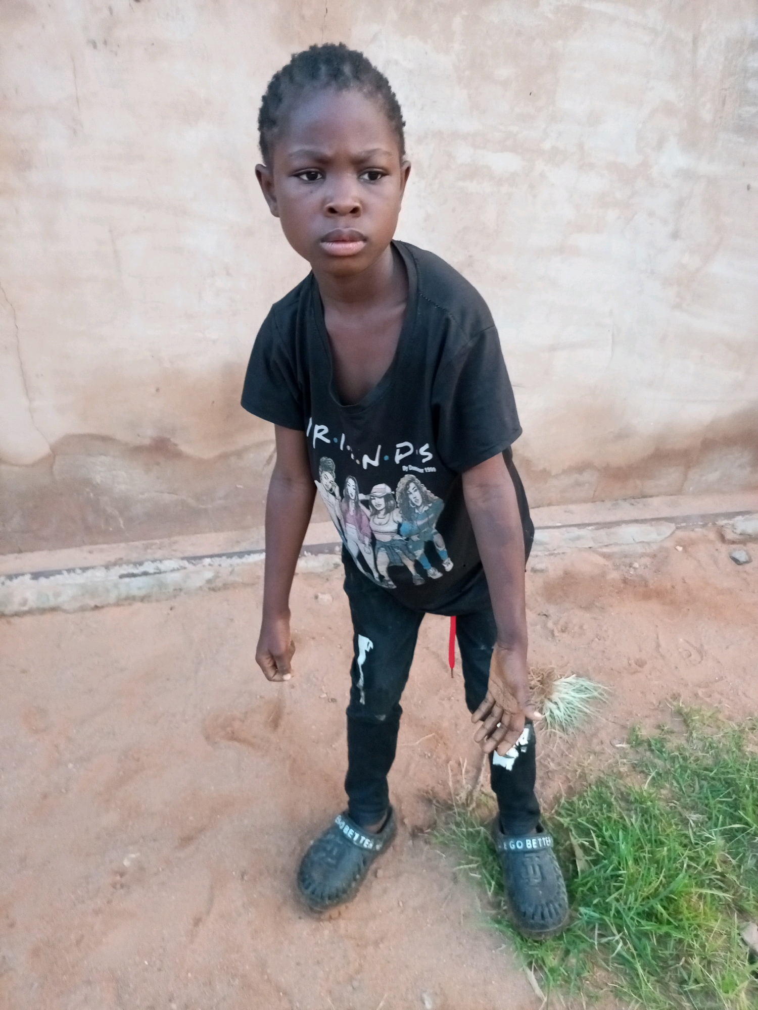 A young Black person, appearing to be a child, stands facing the camera against a neutral, textured wall. They have dark hair styled in short braids and dark eyes. The child is wearing a black t-shirt with a graphic print of cartoon characters and the word "R.I.P.S." printed above them. They are also wearing black pants with a red drawstring visible at the waist, and black clogs with the inscription "GO BETTER" on the strap. The ground in front of the child is composed of sandy dirt with some patches of green grass to the right. The scene is likely outdoors, given the natural lighting and ground texture.