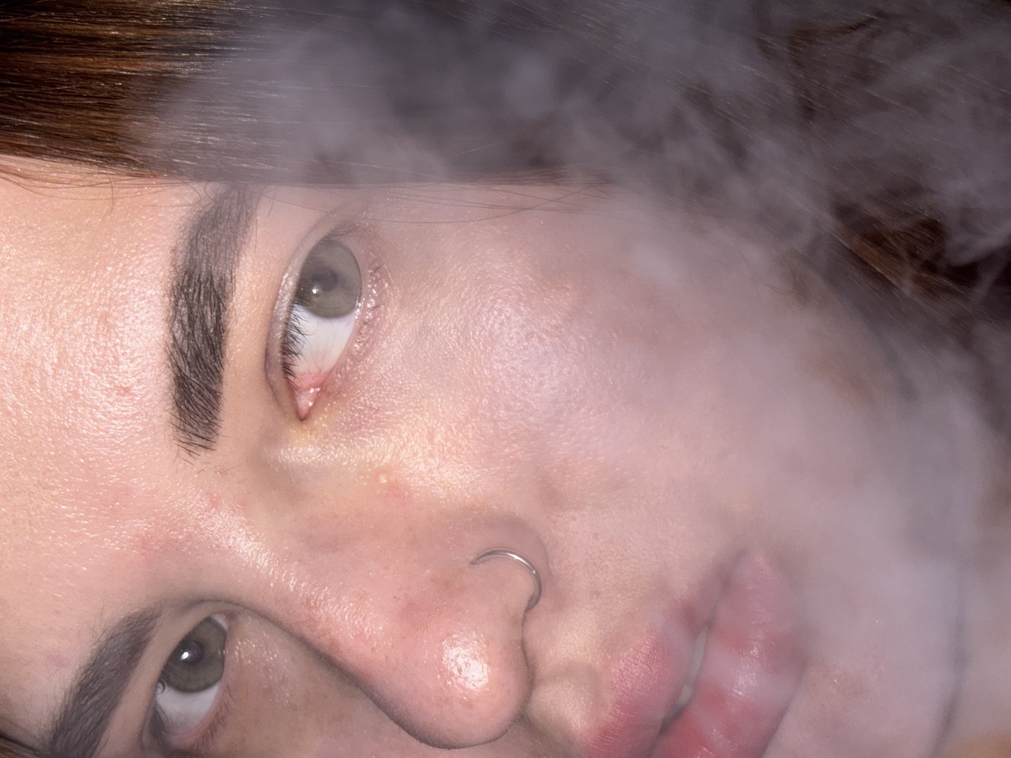 A close-up image shows a person's face partially obscured by smoke or fog. The person has fair skin, brown hair, and light-colored eyes. A silver nose ring is visible. The mouth is slightly open. The overall image is somewhat hazy due to the smoke or fog effect. There is no information to identify this scene as being in Krasnodar, Russia.