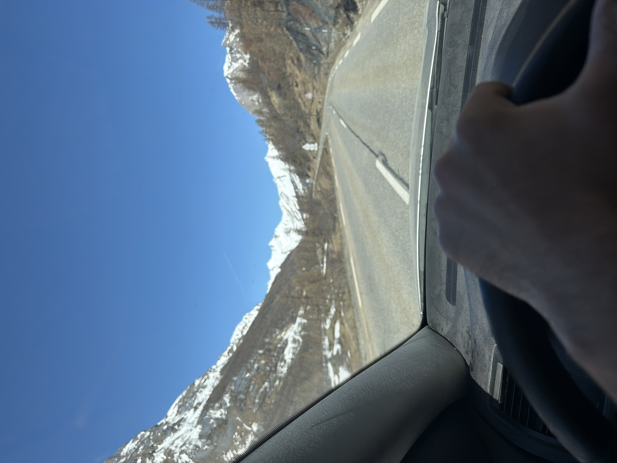 The image is taken from inside a vehicle, likely a car, driving on a paved road. The road winds alongside a mountain range. The mountains are partially covered with snow. The sky is clear and blue. A person's hand and a portion of the steering wheel are visible in the foreground, suggesting that the person is driving. Based on the landscape and context clues, this image was likely taken near Le Monêtier-les-Bains, France.