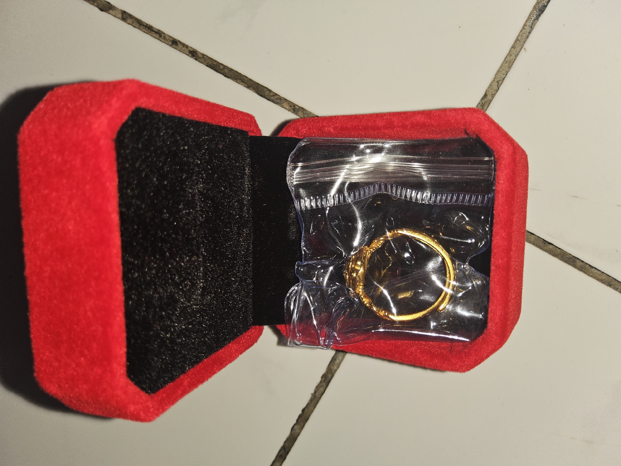 A close-up, overhead shot shows a gold ring, encased in a clear plastic bag, resting inside a red velvet ring box. The ring box is open, revealing a black velvet interior. The box and ring are positioned on a tiled surface. The tiles are off-white with dark grout lines. The lighting creates a shadow in the upper left corner of the image. There are no people visible in the image. The location is not specified in the image or the provided crops, therefore no information regarding the city of Payagaboh, Indonesia can be included.