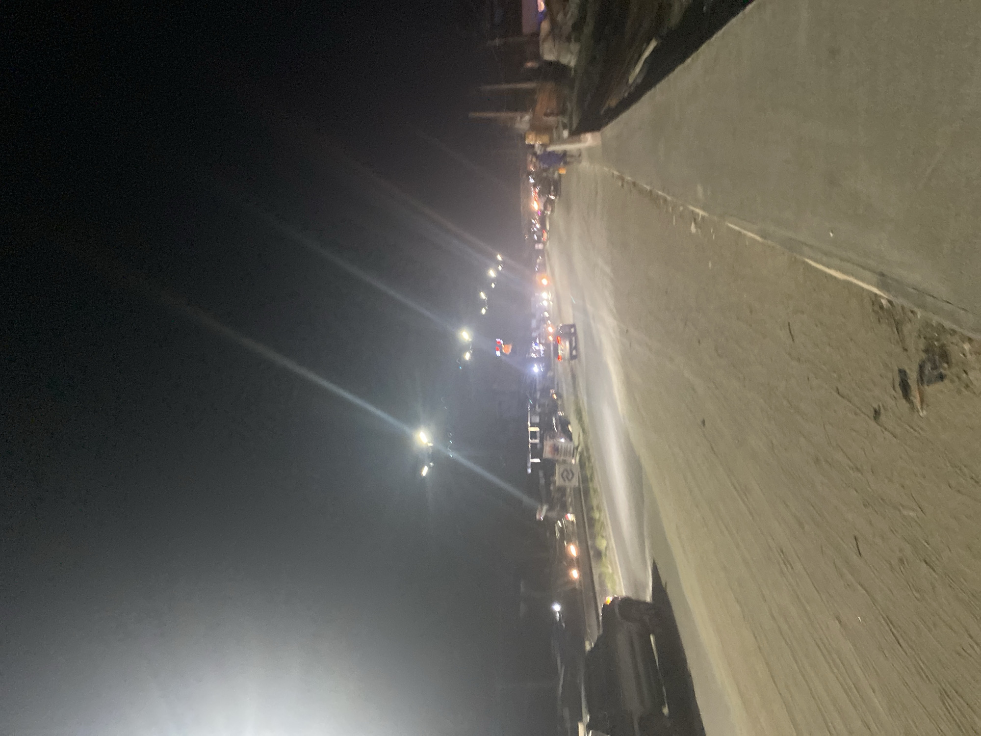 The image depicts a nighttime street scene in an unknown city. A concrete road runs along the right side of the frame. Streetlights illuminate the scene, with light sources appearing along the left side. There are vehicles and what appears to be construction equipment visible in the distance. The sky is dark and devoid of detail. The overall impression is one of a dimly lit urban environment.