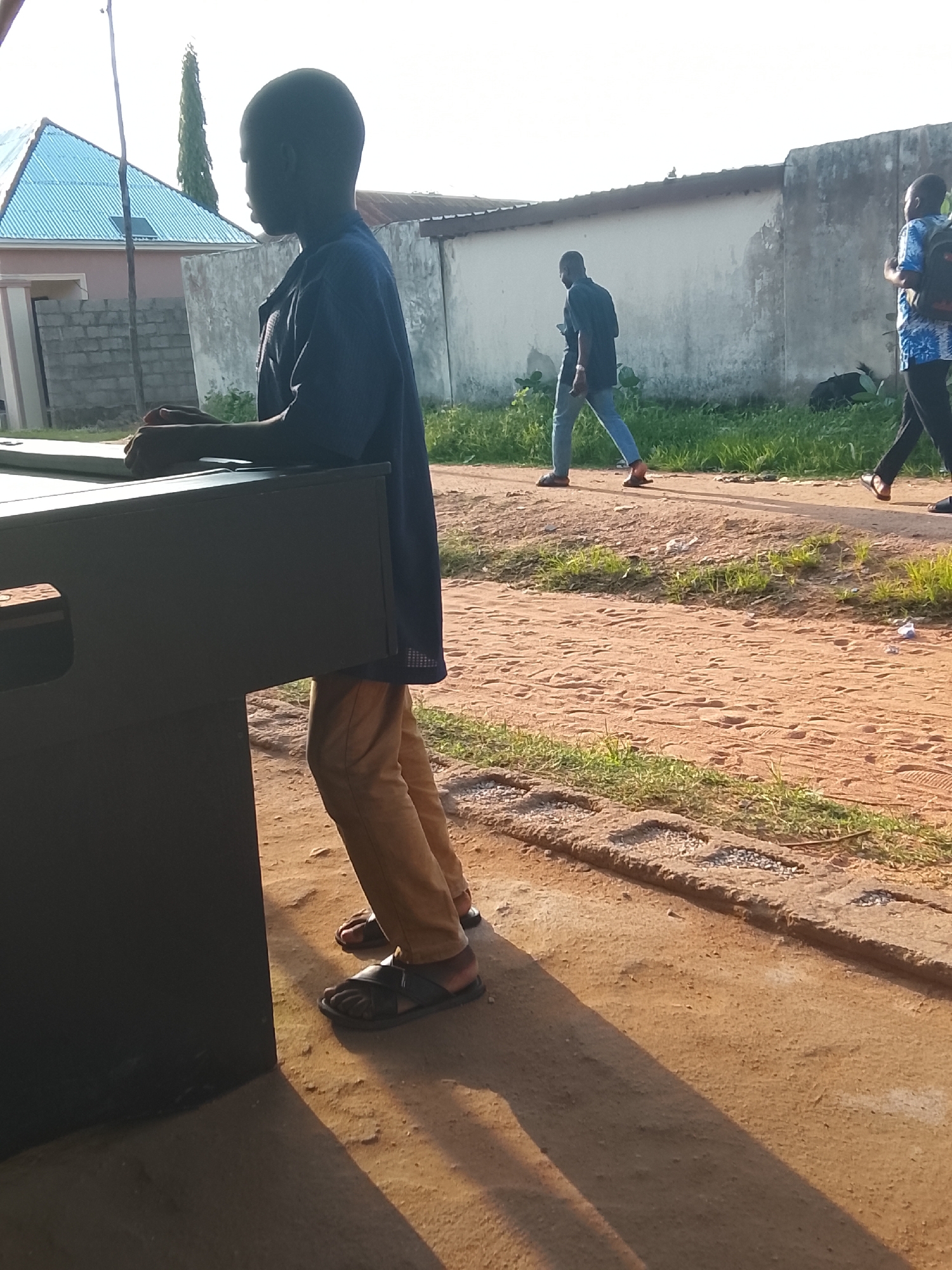 The media file shows an outdoor scene with several people. In the foreground, a young boy is standing next to a dark, rectangular object that appears to be a table or a cabinet. He is wearing a blue shirt and light brown pants, with dark sandals on his feet. His hands are resting on the object. To the right, two men are walking away from the camera. The man closer to the foreground is wearing a blue patterned shirt and dark pants with a backpack. The man further away is wearing a dark shirt and light blue jeans, also with a backpack.
The ground is made of reddish-brown dirt and scattered gravel. There is green grass growing along the side of the path. In the background, there is a building with a blue, corrugated roof and a concrete wall. The scene appears to be set in a somewhat rural or semi-urban environment.
The lighting suggests it is daytime, possibly late afternoon, as long shadows are cast on the ground. The sky is bright, indicating clear weather. There is no visible text in the image. The overall atmosphere is casual and everyday. The location context is Jalingo, Nigeria.