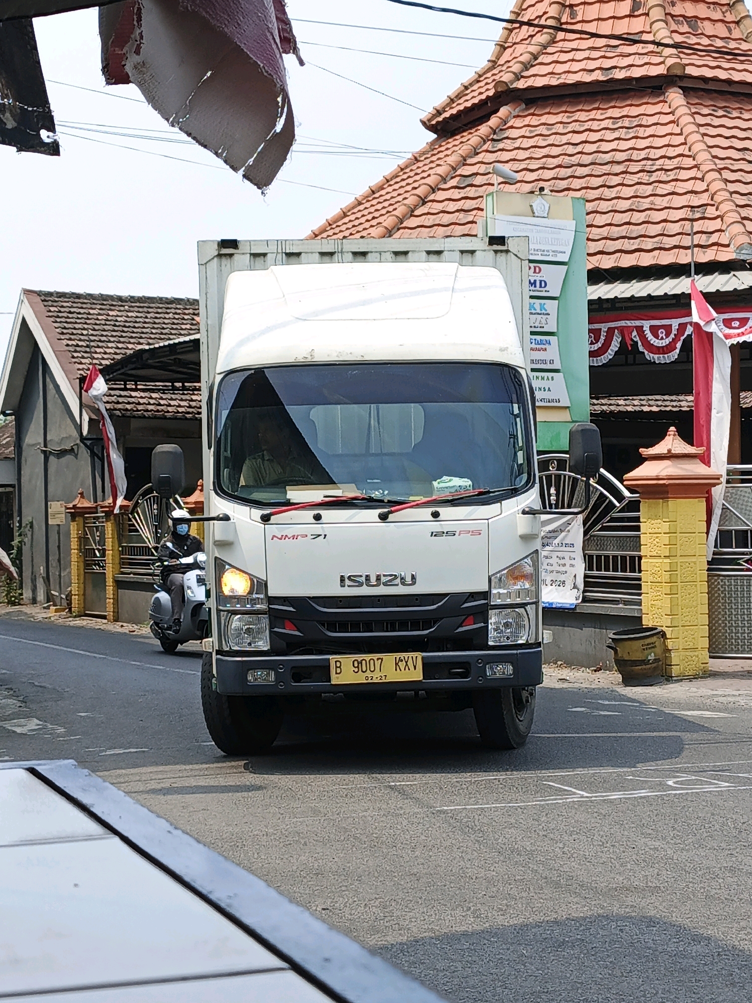A front-view, medium shot captures a white Isuzu NMP71 truck on an asphalt road in Ketegan, Indonesia. The truck's windshield is partially obscured by dangling torn fabric, and a blue scooter with a rider wearing a blue mask and white helmet is visible to the left, partially behind the truck. A building with an orange tiled roof and red and white decorative banners is situated to the right, with a yellow brick pillar in front of it. The road has white lane markings. The sky is overcast.