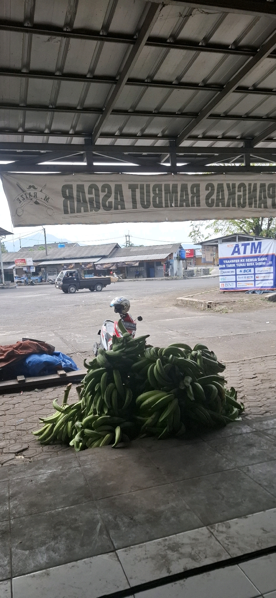 The image depicts an outdoor scene on a street in Sirnagalih, Indonesia. In the foreground, a large pile of green bananas is displayed on a tiled surface. A motorcycle, with a helmet placed on the handlebars, is parked next to the bananas. Behind the bananas and motorcycle, a street with a few vehicles is visible, including a black pickup truck. Several buildings line the street, some appearing to be shops or market stalls. A large banner with Indonesian text and an image of scissors and a comb is hanging above the street. To the right, a white banner advertising ATM services is displayed. The scene is under a covered structure with a metal roof.