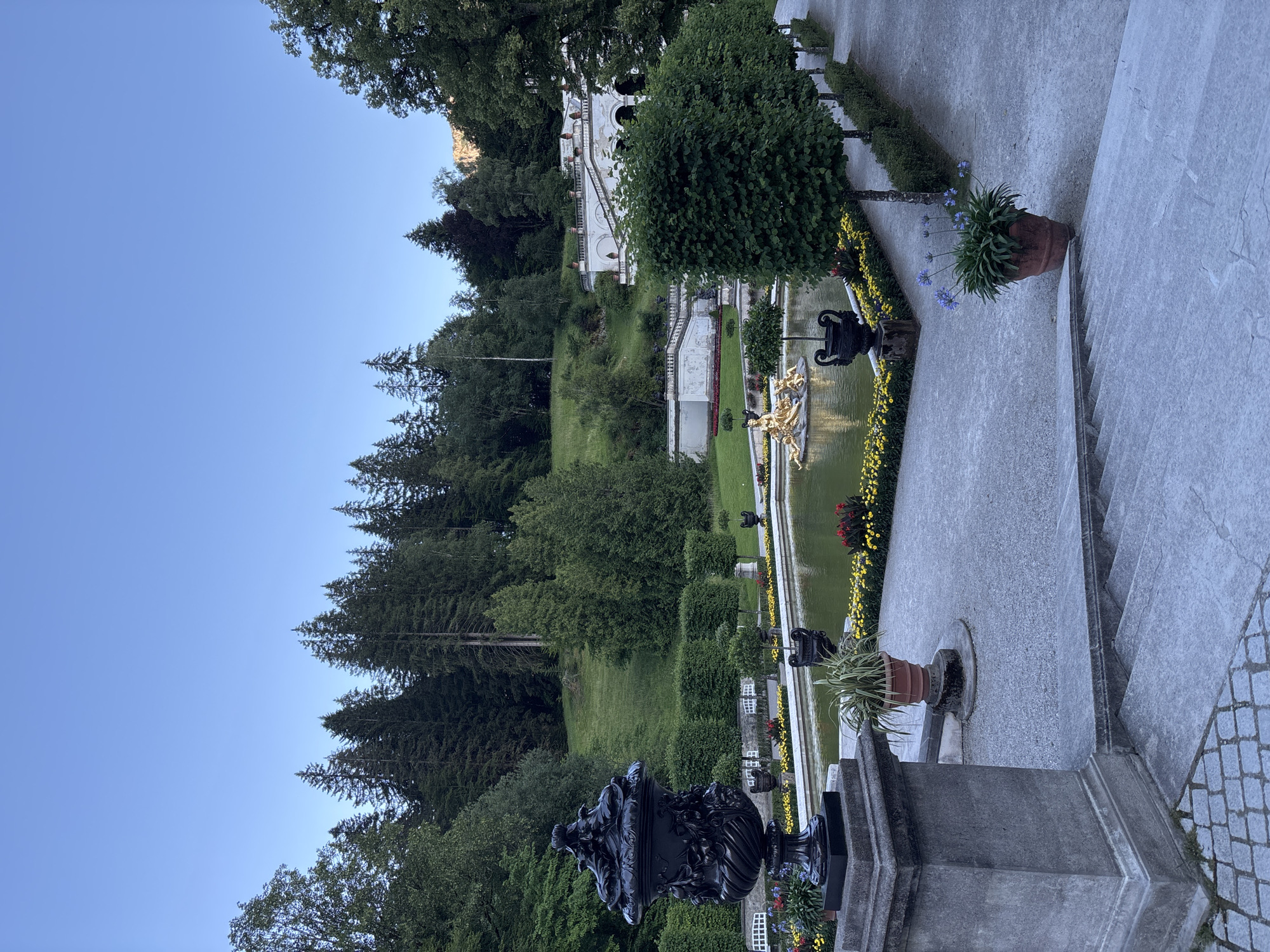 The image depicts a serene garden scene at Linderhof Palace in Ettal, Germany. The foreground features a large, ornate black urn on a stone pedestal, with cobblestone paving at its base. To the right, a set of stone steps leads down towards a pathway lined with meticulously trimmed trees. A terra cotta pot containing blue flowers sits on the gravel path near the steps. Beyond the steps, a rectangular pond with a golden fountain sculpture at its center is visible. The pond is bordered by flowerbeds with yellow and red blooms. In the background, a lush green hillside rises, dotted with trees, and a white palace structure with cascading staircases is nestled against the slope. The sky above is a clear, pale blue, suggesting early morning or late evening. There are no people or animals visible in the image.