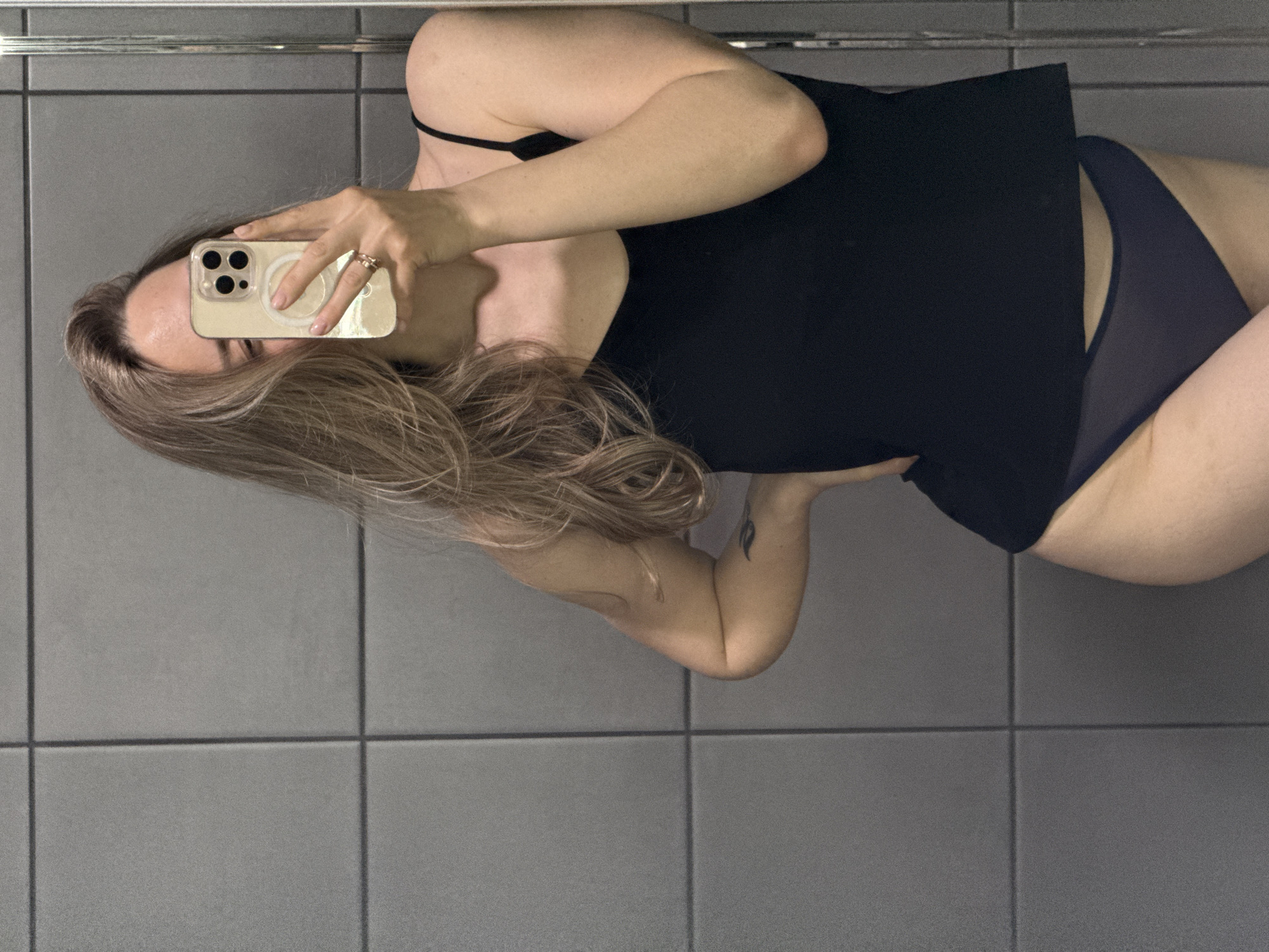 A woman stands in a room with gray tiled floors and walls. She wears a black tank top and dark-colored underwear. She holds a smartphone in front of her face, presumably taking a selfie. She has long, blonde hair. The room is brightly lit. The location is unknown and there is no evidence to suggest the city is Chaikovsky, Russia.