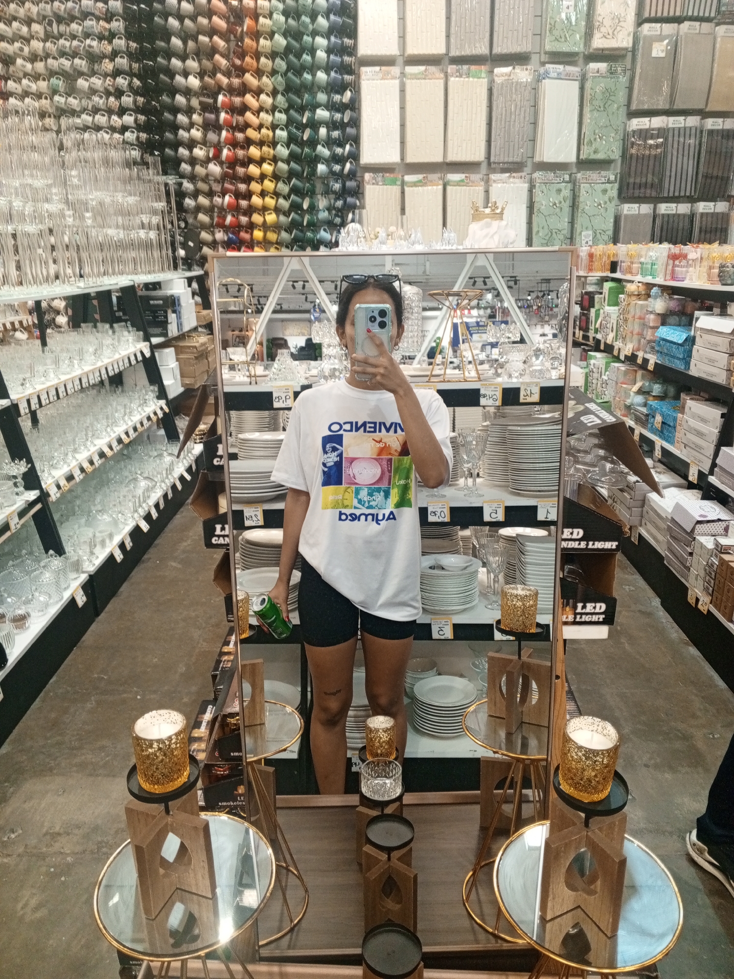 A young woman takes a selfie in a large mirror. The mirror is in a home goods store aisle in Pampatar, Venezuela, surrounded by displays of glassware, plates, candles, and other merchandise. The shelves behind her are full of glasses, mugs, dishes, and decor items. She wears a white t-shirt with a colorful design and black shorts. She holds a green can, seemingly a beverage. The mirror is flanked by small tables decorated with gold votive candles.
