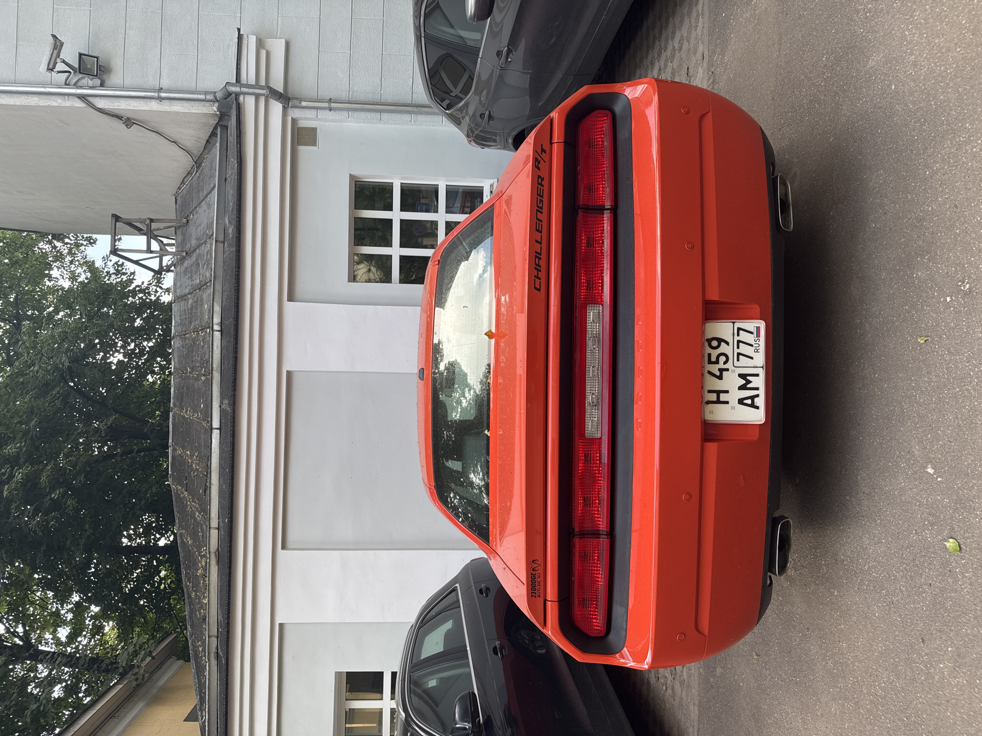 The rear view of a bright orange Dodge Challenger R/T is captured from a low angle, parked on asphalt. The car's taillights are prominent, and a license plate with Russian characters (H 459 AM 777) is affixed. The background features a grey and white building with multiple windows, a section of a dark-colored car on the left, and a lighter grey SUV on the right. Overhead, trees with lush green foliage are visible against a cloudy sky. The scene is set in a city environment, likely Moscow, Russia, given the license plate and architectural style. There are no people or animals present in the image.