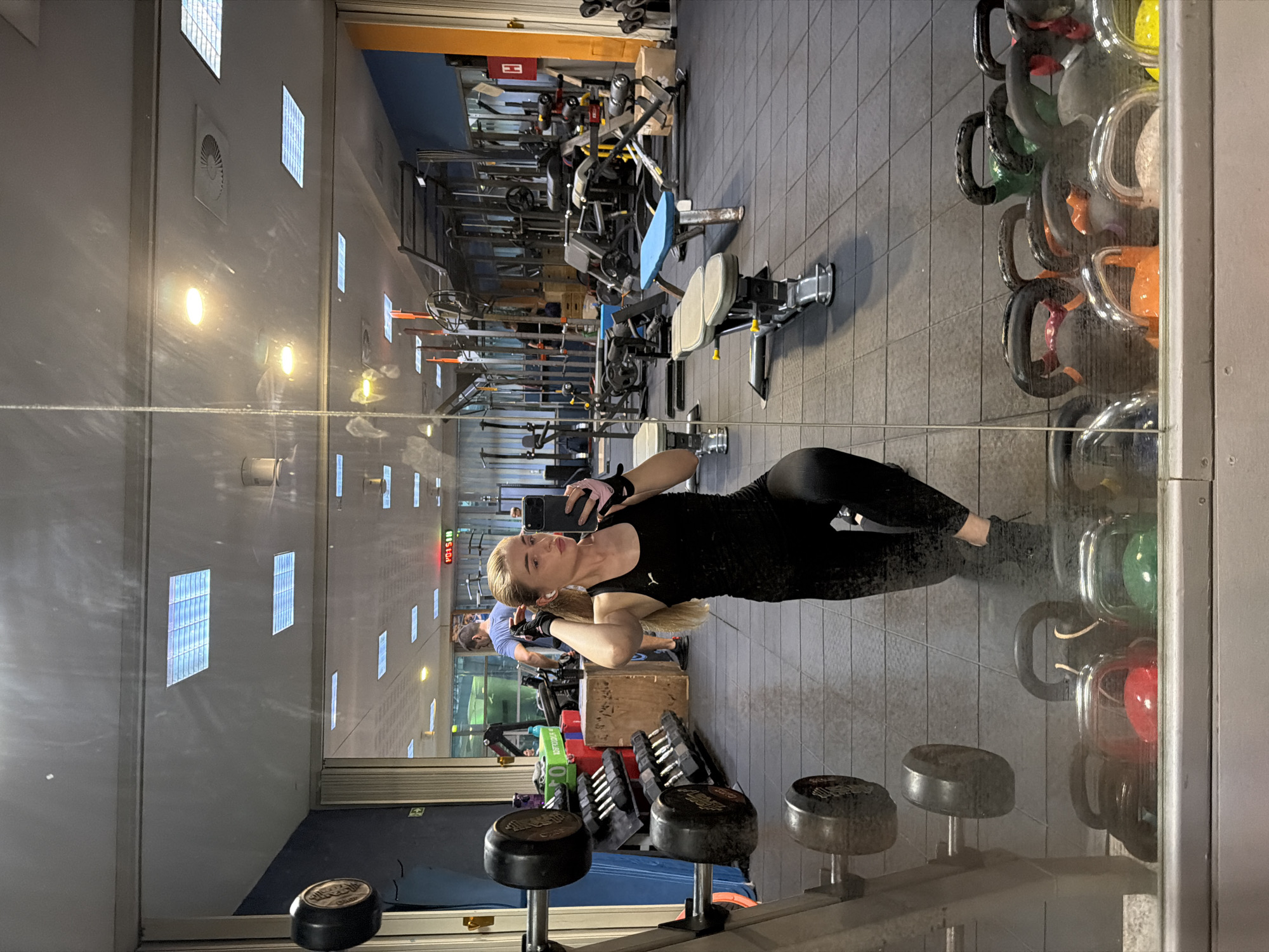 The image shows a young woman with blonde hair, wearing a black athletic outfit, taking a mirror selfie in a gym. She is holding her phone in her right hand and adjusting her hair with her left hand, wearing earbuds and black gloves with pink accents. She is in a squatting position. The gym is equipped with various weightlifting equipment, including dumbbells on racks, weight benches, and other machines visible in the reflection. A digital clock displaying "13:51:08" is mounted on the wall in the background. Another person, a man wearing a blue t-shirt, is visible in the reflection, standing and looking away. The floor is covered with dark grey rubber tiles. In the foreground, reflected in the mirror, are several kettlebells and other weights. The lighting suggests an indoor environment, likely during the daytime.
