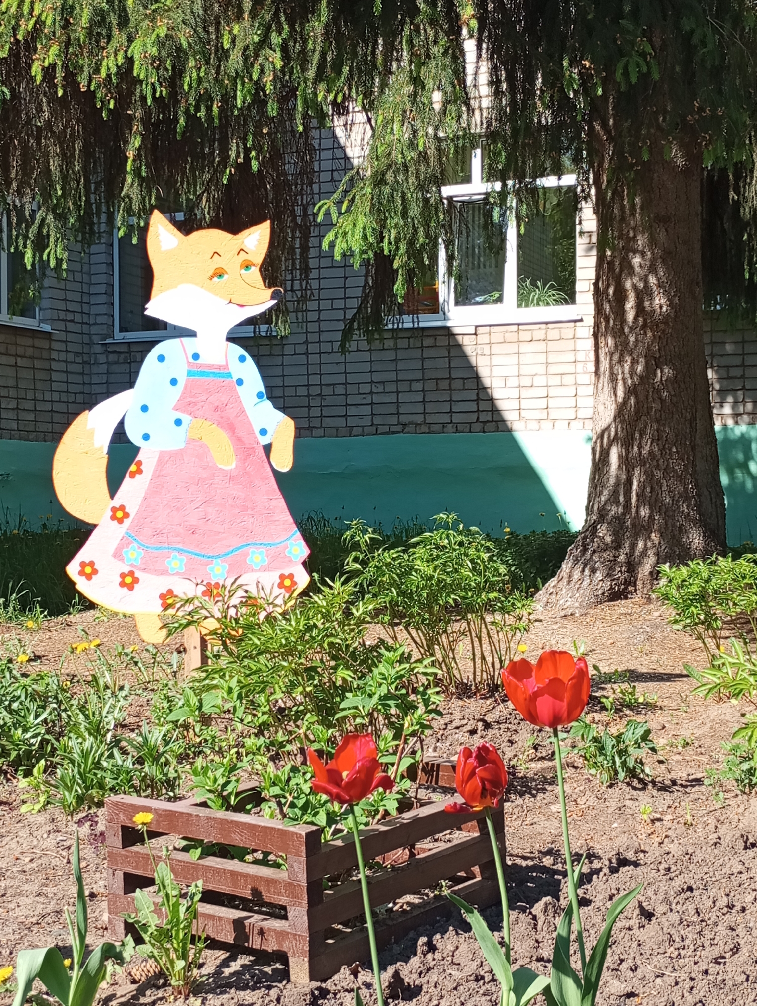 The image shows a garden scene in Izhevsk, Russia. A painted, cartoon-like fox figure stands in the garden. The fox wears a pink apron with flower accents and a blue dress with white sleeves and blue polka dots. The garden contains a small, dark brown, rectangular wooden planter box. Several red tulips are in bloom, interspersed among other green plants and weeds. A large evergreen tree is behind the fox and garden, next to a light brick building with a white-framed window. The scene appears to be well-lit by sunlight.