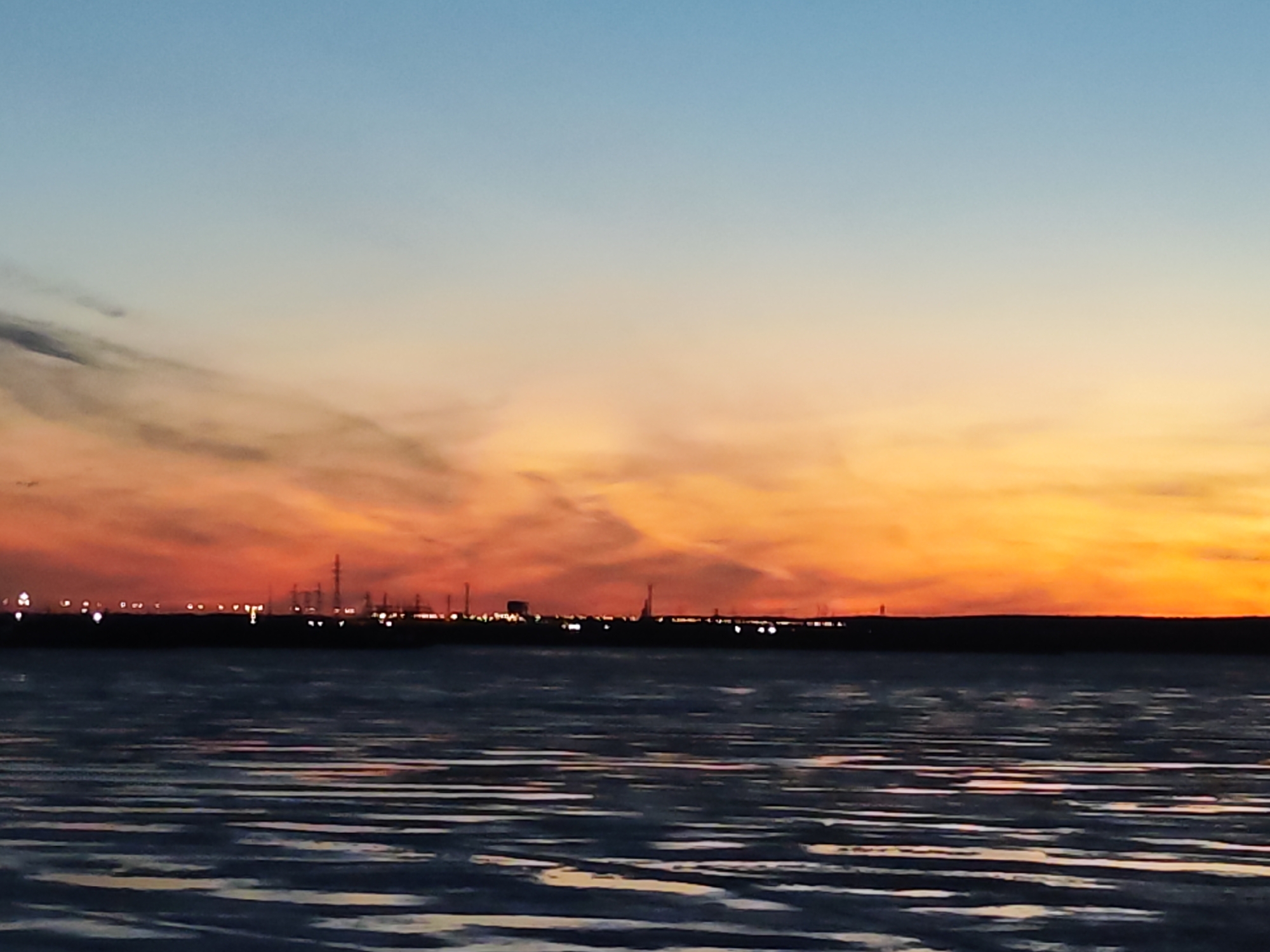 The image captures a twilight scene over a body of water. The sky transitions from a light blue at the top to vibrant orange and red hues near the horizon. The water in the foreground displays prominent horizontal patterns of light and shadow, possibly due to ripples or tidal effects. In the distance, a dark silhouette of a landmass is visible, punctuated by scattered lights that suggest a town or industrial area. The location is Chaikovsky, Russia. The scene evokes a sense of calm and solitude.