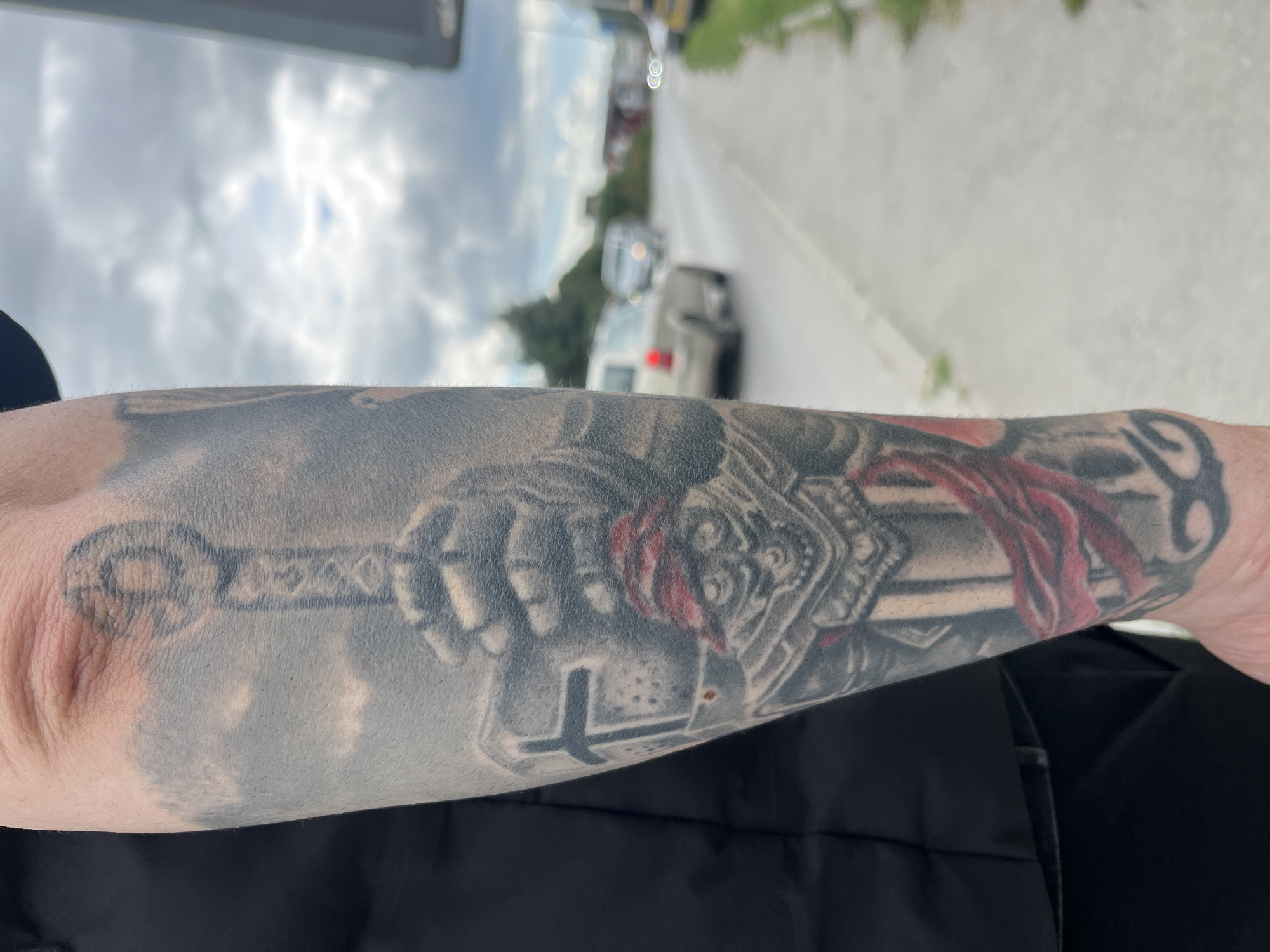 The image shows a person's left forearm with a tattoo depicting a medieval knight holding a sword. A red cloth is draped around the knight's head or helmet. The background is blurry and shows a road with cars and greenery, possibly taken outdoors on a cloudy day. Without further information or visual cues, it's impossible to determine if the location is Varna, Bulgaria.