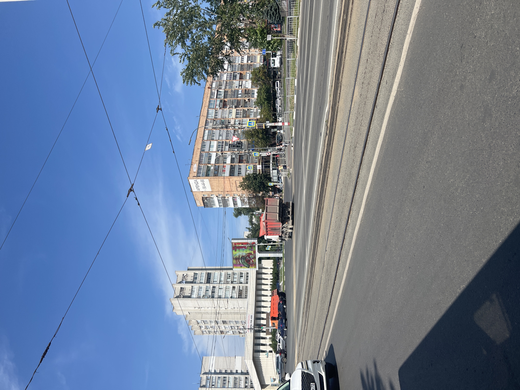 This is a street-level, medium-shot, eye-level photograph of a wide, multi-lane paved road with tram tracks in the foreground. The road surface is asphalt and appears worn in places. The sun is shining brightly, casting a shadow from the left edge of the frame onto the road. In the middle ground, there is a complex intersection with pedestrian crossings, traffic lights, and several vehicles. A red dump truck is visible moving through the intersection. To the left of the intersection, there are several multi-story, Soviet-era apartment buildings. One building on the far left has a white facade and is connected to a lower, wider building with a sloping roof. Another taller apartment building to its right is light grey. In the center of the image, a large, brightly colored digital billboard is mounted on a pole. To the right of the intersection, there is a prominent, large, orange-brick apartment building with many balconies. The building has a sign that reads "АРЕНДА 98-44-86". A large, mature tree with green foliage is to the right of this building, partially obscuring it. Overhead, numerous power or tram wires crisscross the bright blue sky, which has a few wispy clouds. The scene is set in the city of Ulyanovsk, Russia.