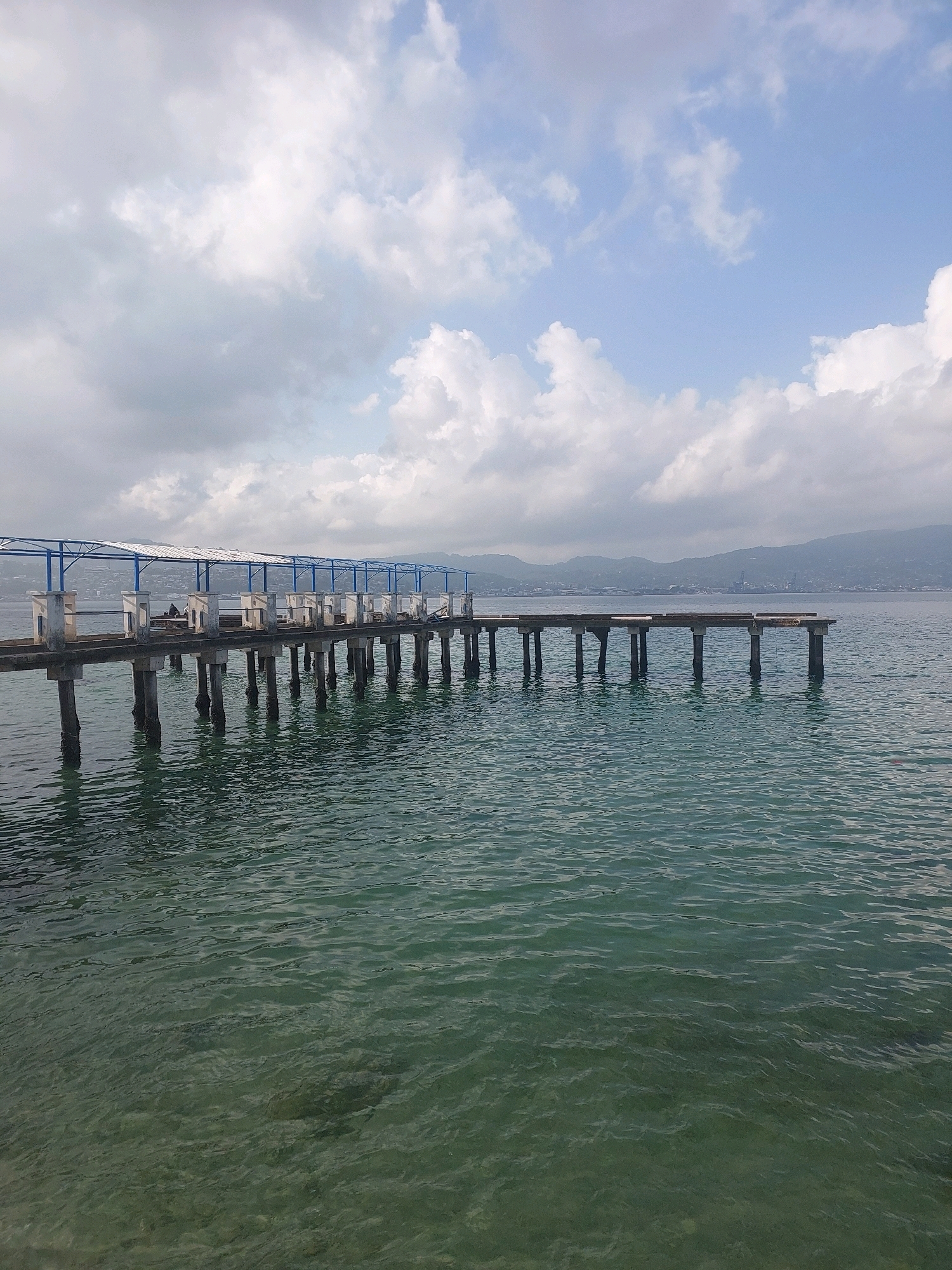 The image depicts a pier extending into the turquoise waters of Ambon, Indonesia. The pier, constructed with concrete pillars and a walkway, is partially covered by a blue-framed canopy with a white roof. The sea is calm, with gentle ripples reflecting the cloudy sky above. In the distance, the coastline is visible, with hills and a town nestled along the shore. The overall atmosphere is serene and quiet, suggesting a peaceful day at the waterfront. There are no visible people on the pier, but there appears to be a person sitting on the pier.