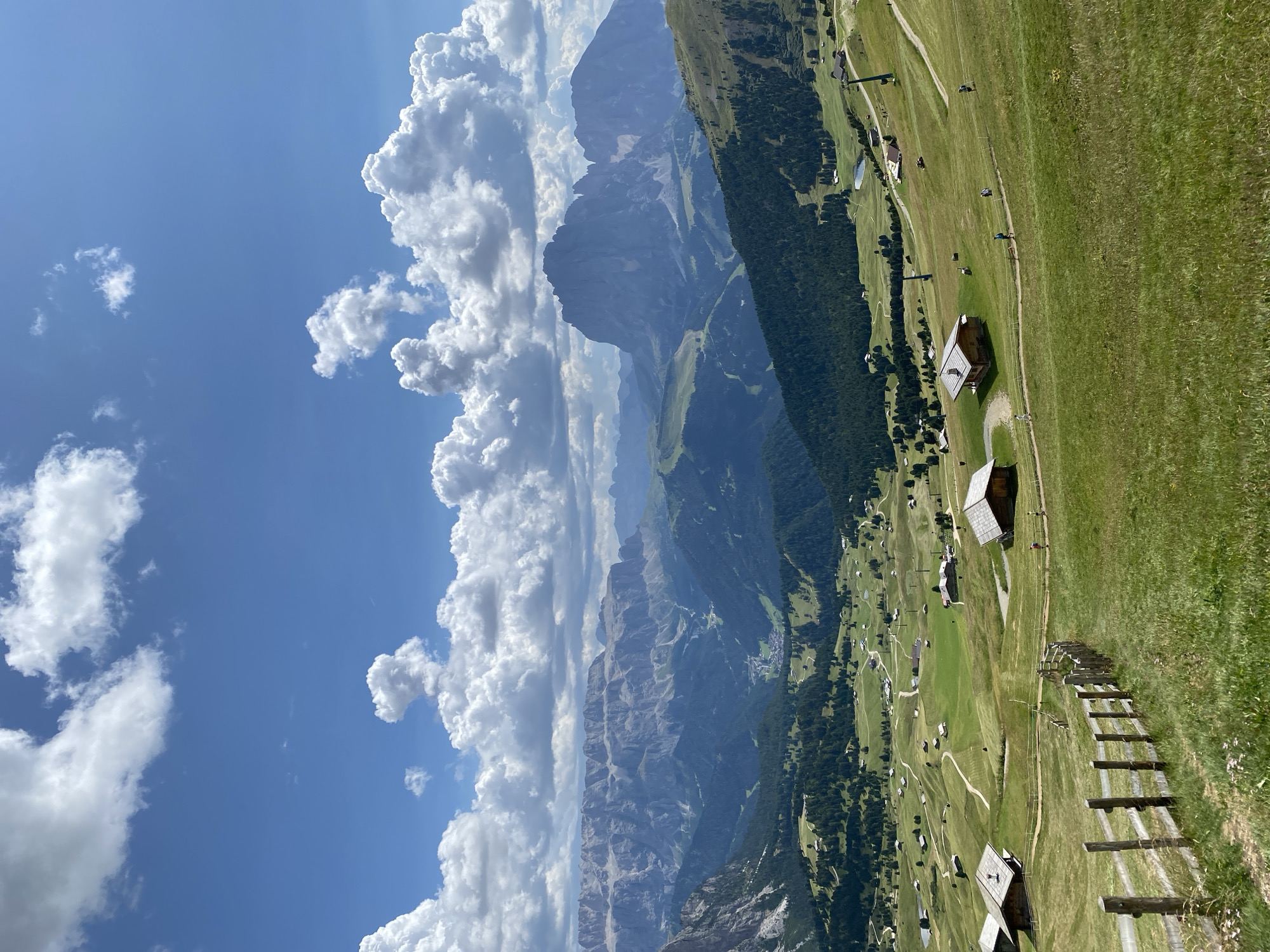 This is a scenic, elevated, outdoor, daytime, long-shot photograph of a mountainous landscape in the Italian Dolomites. The sky is blue with scattered white cumulus clouds. In the foreground, a wooden fence with three horizontal planks runs diagonally from the lower left corner towards the center. Beyond the fence, a lush green meadow slopes down, dotted with several small, rustic wooden buildings with dark pitched roofs. A winding dirt path meanders through the meadow, with a few distant figures walking on it. Further in the midground, more clusters of wooden structures and a small body of water are visible, nestled amidst rolling green hills and dark green pine forests. In the background, imposing, rugged mountain ranges stretch across the horizon, their gray, rocky faces partially obscured by haze and the shadows of the clouds. The overall impression is one of a tranquil, rural alpine setting.