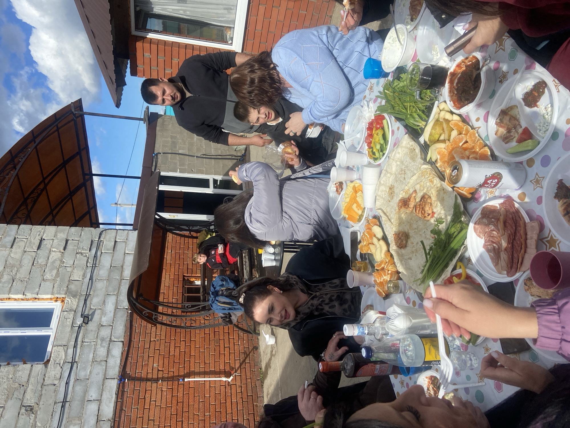 People are gathered around a table outdoors in Ольховка, Russia, eating a meal. The table is covered with a patterned tablecloth and various dishes of food, including meats, vegetables, fruits, and bread. There are also drinks, disposable plates, cups, and plastic utensils. People are holding food or utensils, and some appear to be talking or laughing. A brick building is in the background, along with a covered structure and some greenery. The sky is visible with clouds.