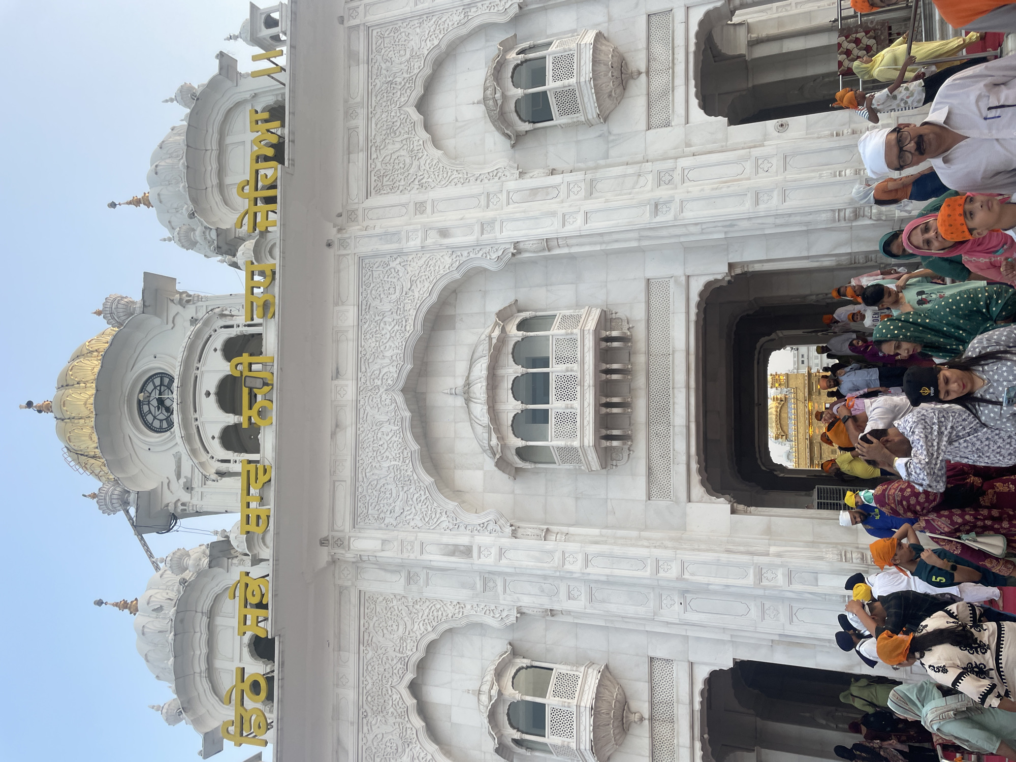 The image shows the Darshani Deodi, the main gateway to the Harmandir Sahib (Golden Temple) in Amritsar, India. The structure is made of white marble with intricate carvings. Yellow lettering in Gurmukhi script is visible above the entrance. A large clock is mounted on the facade of the building. A crowd of people, many wearing turbans and colorful clothing, are entering or gathered around the gateway. Some individuals are looking at the camera, while others are engaged in conversation or moving through the area. The sky is clear and light blue. The scene suggests a busy day at a significant religious site.