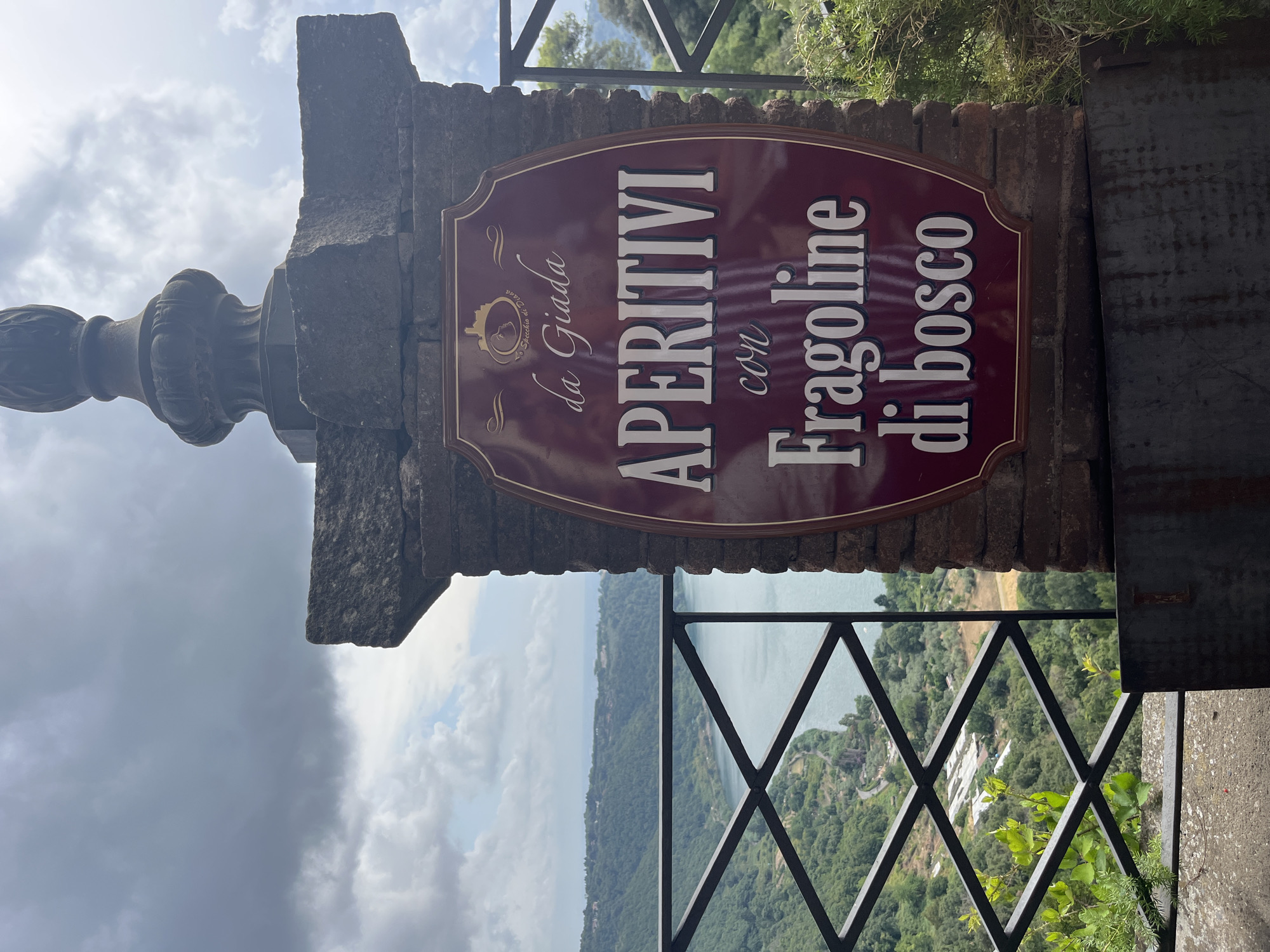 A close-up, eye-level, outdoor, vertical shot captures a burgundy sign with gold lettering attached to a stone pillar. The sign reads "da Giada APERITIVI con Fragoline di bosco," with a small illustration of a castle and a woman's profile at the top. Behind the sign and pillar, a dark metal railing encloses an overlook. Beyond the railing, a picturesque view of Nemi, Italy, unfolds. A serene lake, Lake Nemi, is visible in the distance, surrounded by lush green hills dotted with trees and some buildings. The sky above is overcast with dramatic clouds. The pillar is made of weathered brick and stone, topped with an ornate dark metal finial. The overall scene suggests a scenic spot offering aperitifs with a view.