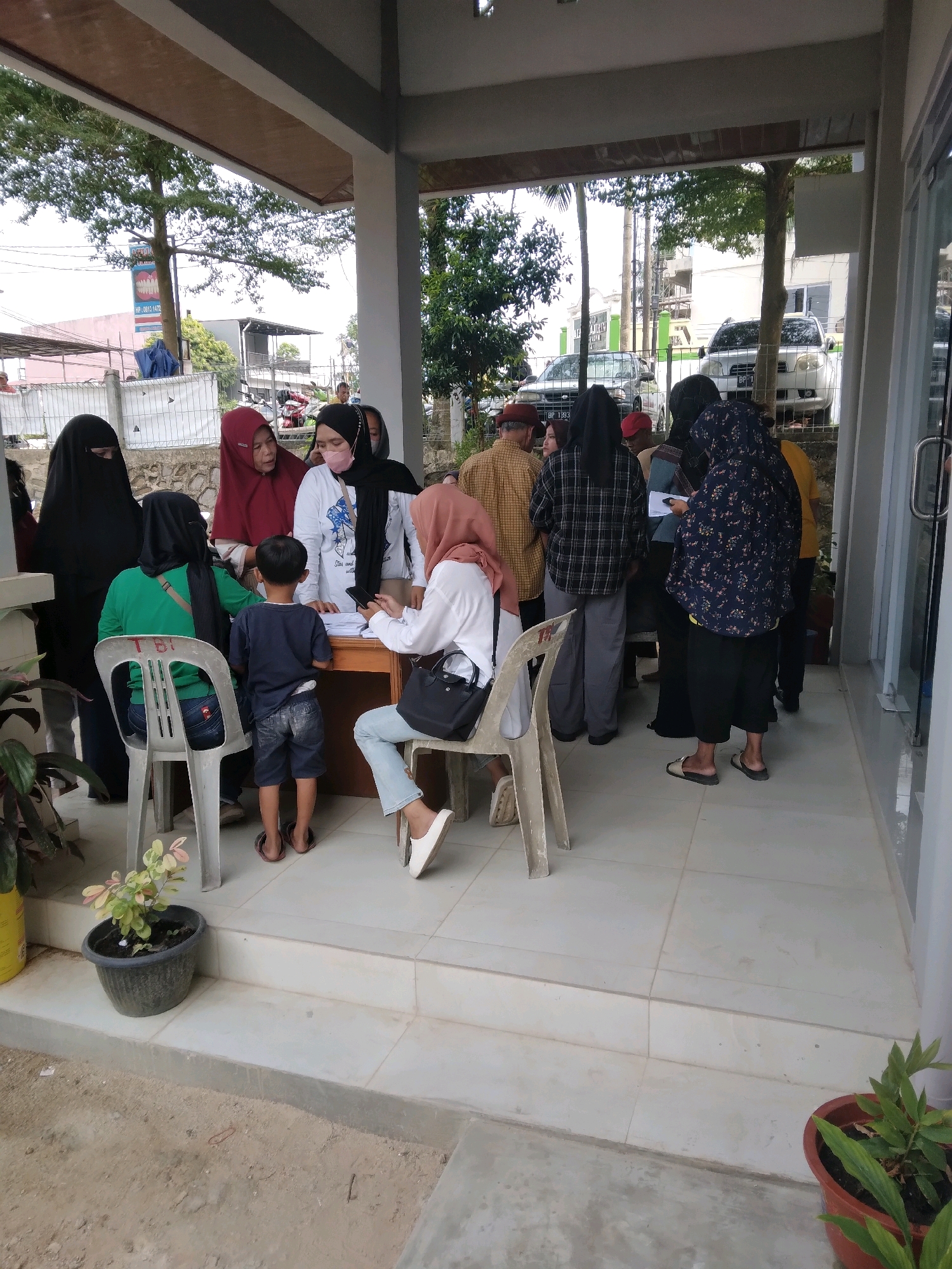 The image depicts a scene outside a building, likely a government office or a public service facility, in Batam, Indonesia. Several people, predominantly women dressed in hijabs and modest clothing, are gathered. Some are standing, while others are seated on plastic chairs. A wooden table is present, with papers on it, suggesting a registration or service counter. One woman, wearing a white shirt and a pink face mask, appears to be attending to some paperwork or assisting individuals. A young boy is standing near the table, facing away from the camera. The environment appears to be an outdoor waiting area or a covered walkway, with tiled flooring and a concrete ground. Trees and buildings are visible in the background, along with parked vehicles, indicating an urban or semi-urban setting. The overall impression is one of a queue or a waiting line for a service.