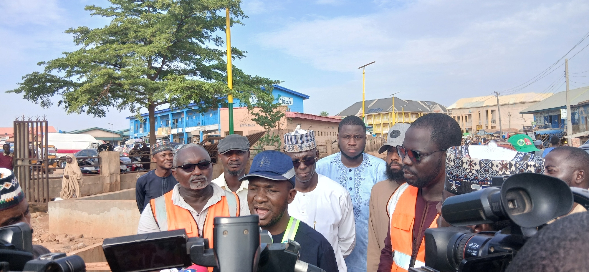 The image depicts a group of men gathered outdoors in a public space, likely a street or market area, in Zaria, Nigeria. The foreground is dominated by two professional video cameras, with one person in an orange safety vest speaking into a microphone that is part of the camera setup. Several other men are visible behind the cameras, some looking towards the speaker, others at the cameras, and some have their faces obscured.
The men are dressed in a mix of casual attire and traditional Nigerian clothing, including embroidered shirts and caps. One man in the center is wearing a light blue, intricately embroidered shirt and a dark cap. Another man to his left is wearing a white shirt and a patterned cap. The man in the orange vest has a blue baseball cap on and appears to be the focus of the recording.
In the background, there are buildings, including a prominent blue building with the word "Peak" visible, suggesting a commercial area. There are also trees, a street light, and what appears to be parked vehicles and market stalls. The overall atmosphere suggests a news gathering or public event. The sky is partly cloudy, indicating daylight conditions. The ground appears to be unpaved in some areas.