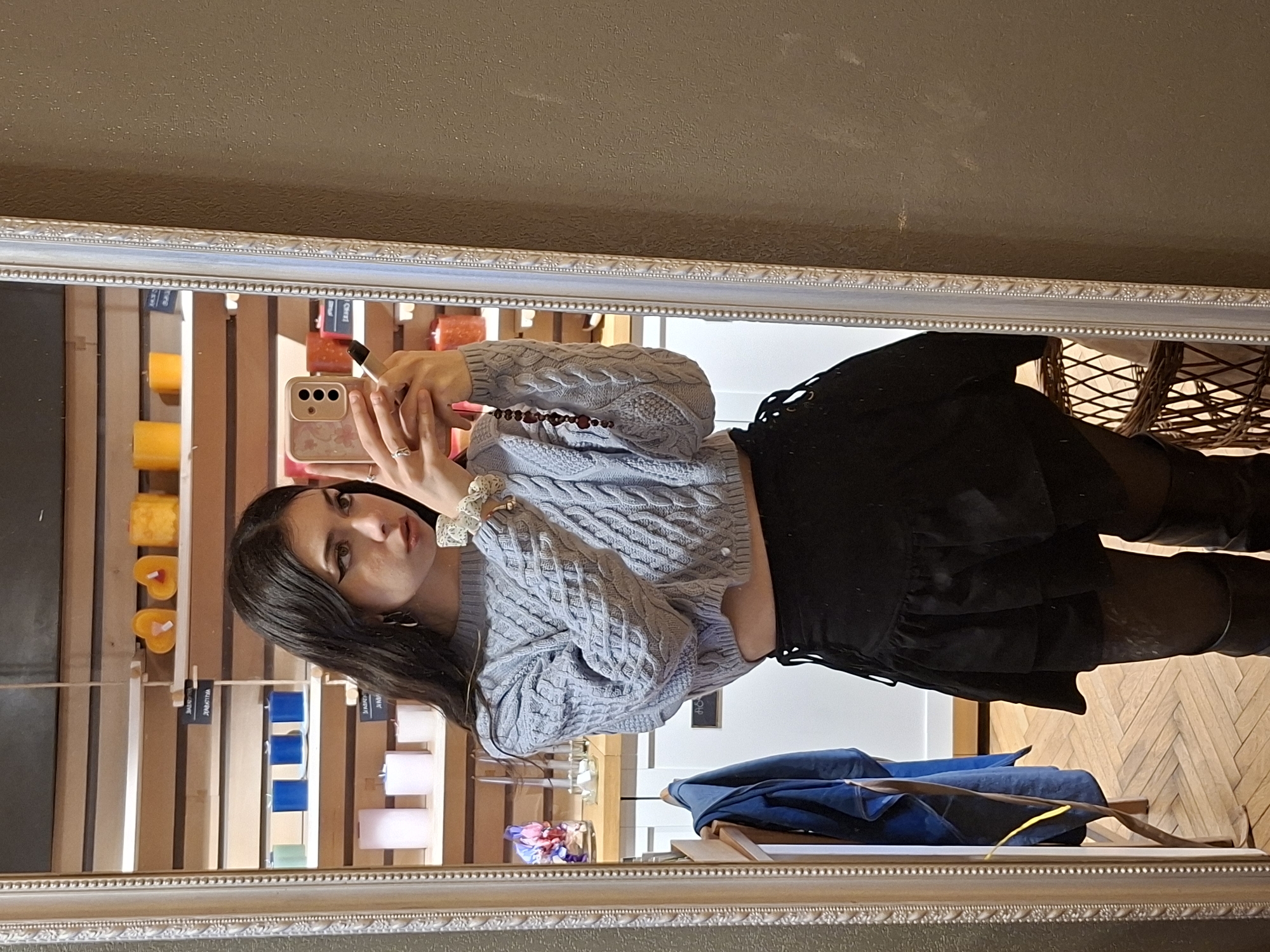 A young woman is seen taking a selfie in a mirror within a shop. She is wearing a light blue cable-knit sweater, a black skirt, and black tights or leggings. She is holding a smartphone in her right hand, and a lipstick in her left. The reflection in the mirror shows candles of different colors. The woman appears to be located indoors near an assortment of goods for sale, possibly in a shop in Sofia, Bulgaria.