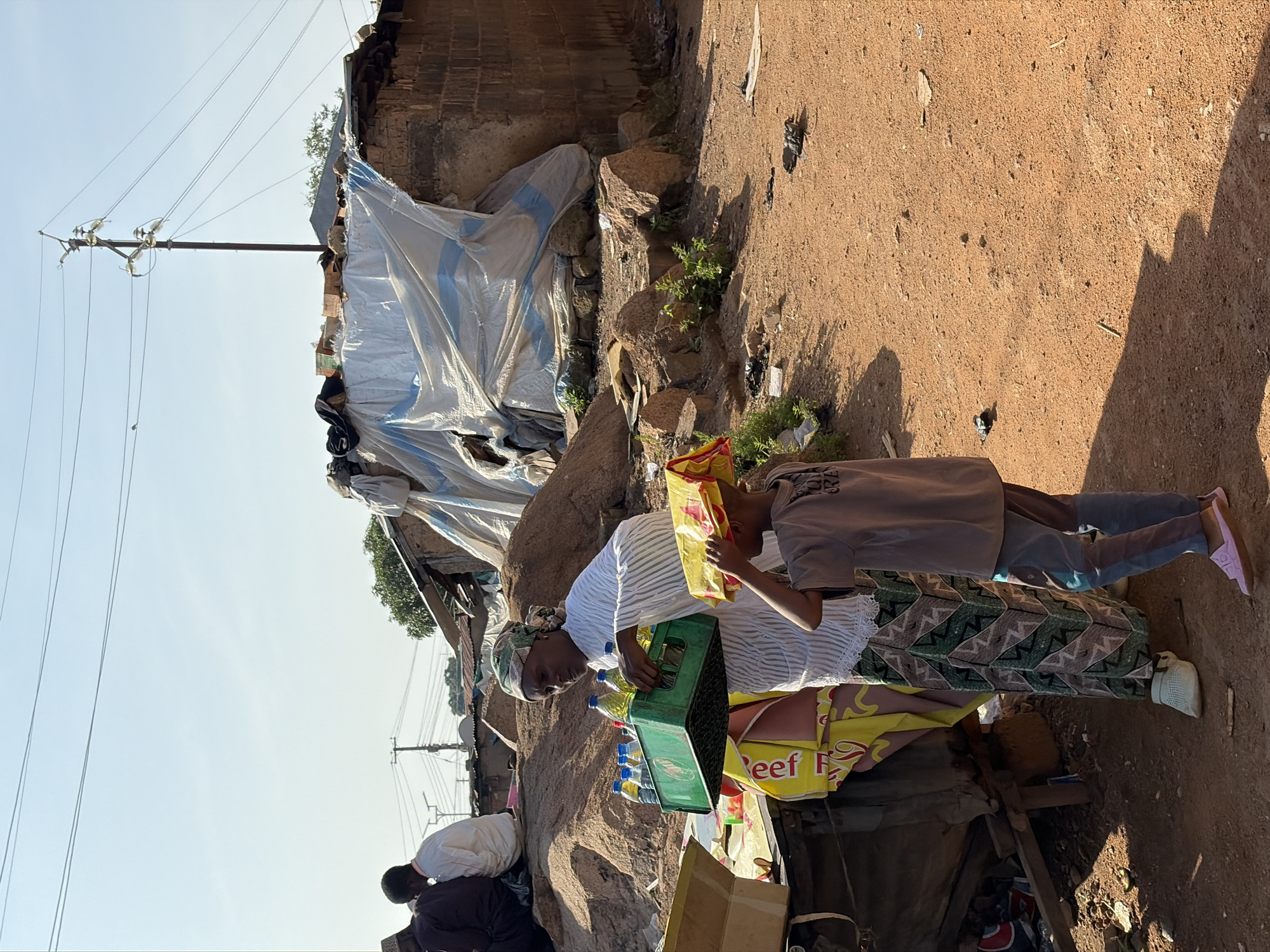 In Jos, Nigeria, a woman wearing a headscarf and a white striped top stands in front of a makeshift market stall. She is holding a green plastic crate filled with bottled drinks. Beside her, a young boy, wearing a grey t-shirt and blue and grey striped pants, holds a yellow and red plastic bag over his head. In the background, a structure with a white and blue tarp draped over it is visible, along with some rocky terrain and a utility pole with power lines. The ground is dusty and reddish-brown, suggesting a dry, arid environment. Other people can be seen in the distance, some seated. The scene depicts a moment of commerce or daily life in what appears to be an informal settlement or market area.