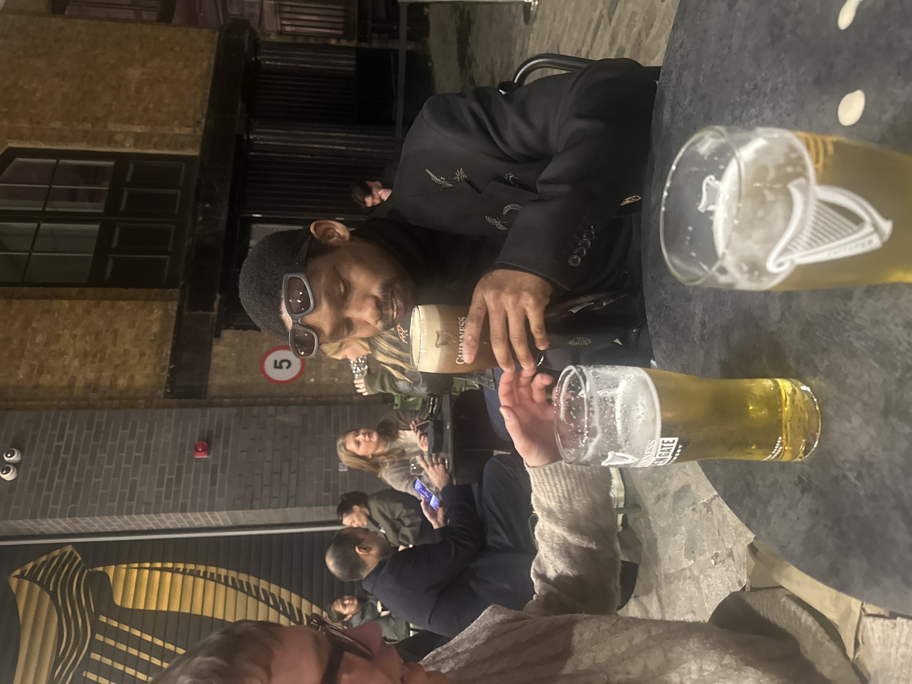 The image captures a vibrant nighttime scene at what appears to be a pub or brewery patio, filled with people socializing. The photograph is oriented vertically, showing the scene from a slightly elevated perspective looking down at a table.
In the immediate foreground, a person with dark skin and short, curly dark hair is prominently featured. They are wearing dark-framed sunglasses and a stylish black jacket or blazer adorned with intricate, light-colored (possibly silver or white) embroidered designs, resembling birds or abstract patterns, on the chest and sleeves. They are holding a glass of dark stout beer, identifiable by the "GUINNESS" text visible on the glass, and appear to be smiling or laughing as they look downwards and slightly to their left. Their left hand rests on the dark-colored, round table.
On the table in front of them, two other glasses are visible, both containing lighter-colored lager or ale. One of these glasses clearly displays the iconic harp logo associated with Guinness. The other lighter beer glass has text partially visible, which reads "GUINNESS OPEN GATE BREWERY".
In the middle ground, several other patrons are visible. A person with lighter-colored hair is seated behind the central figure, looking towards the left and holding a smartphone. Further back, another individual with dark hair, wearing a dark jacket, is also seated. Additional blurry figures populate the background, suggesting a bustling atmosphere.
The setting features brick walls, predominantly dark gray or brown, with dark-framed windows or glass panels. On one of the brick walls, a round red sign with the white number "5" is clearly visible. Below this, a large, golden or bronze mural or art installation with wavy, striped patterns is seen, partially obscured by the people. This mural also hints at the Guinness brand, possibly stylized waves or a harp. Mounted higher on the brick wall are two small, white, circular objects (possibly security cameras or lights) and a small red rectangular box, which could be a fire alarm.
The scene is illuminated by artificial light, indicating that it is evening or night. The overall mood appears convivial, with people enjoying drinks and company.