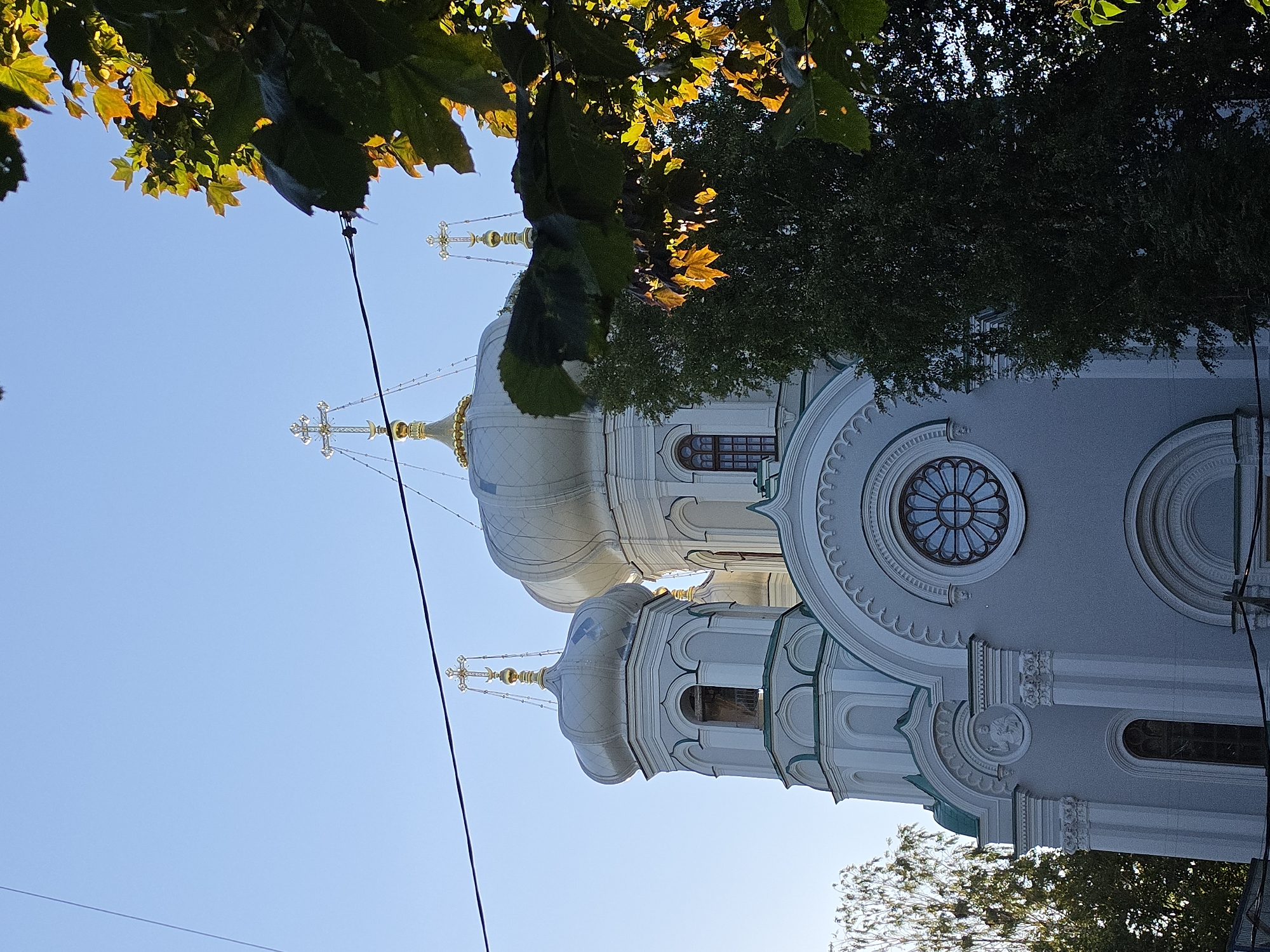 An exterior, low-angle shot captures the domes and facade of a pale grey Orthodox church, partially obscured by lush green foliage and tree branches in the foreground. The sky above is a clear, bright blue. The church features multiple domes, the largest of which is prominent in the center, topped with a golden cross. Smaller domes and towers with similar golden crosses are visible to the left and behind the main dome. The facade exhibits architectural details such as arched windows, decorative molding, and a circular stained-glass window above a smaller arched entrance. A wire runs horizontally across the upper portion of the image. The overall scene suggests a sunny day. This is the Church of the Intercession of the Most Holy Theotokos in Gatchina, Russia.