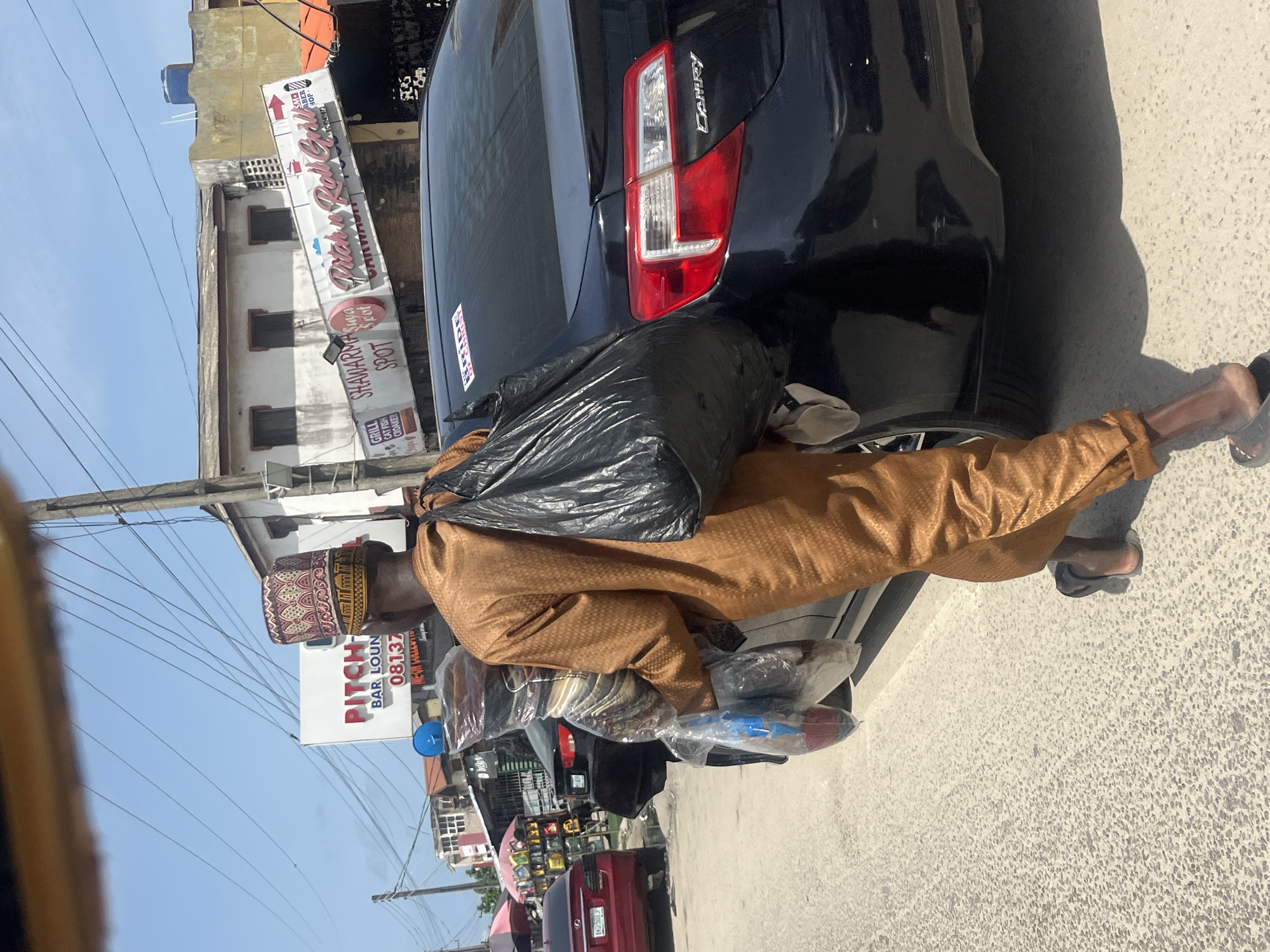 A man dressed in a brown traditional robe and a patterned cap is leaning against the trunk of a dark-colored Camry sedan. He has placed a large black plastic bag on the trunk. He is also carrying wrapped items. The car is parked on a street in Ado, Nigeria. Visible in the background are storefronts with signs for "Quick Roll Grill," "SHAWARMA Suya SPOT Spot," and "PITCH BAR LOUN." A red car is partially visible in the lower background. The street appears to be paved, and there are utility poles and wires overhead. The license plate of the red car is AKD-501 JP.