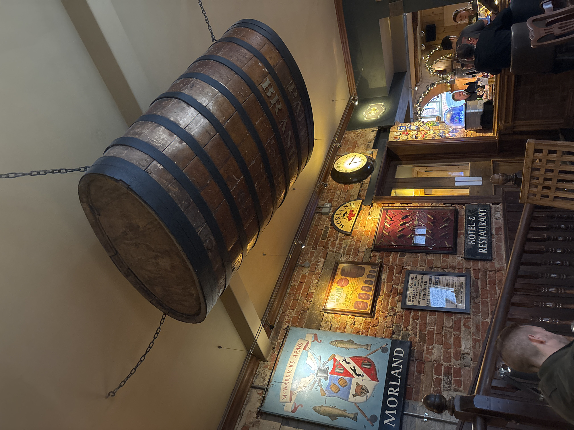 The image shows the interior of a pub in the Vale of White Horse, United Kingdom. A large, wooden barrel hangs from the ceiling by chains. On the brick wall are various signs and framed pictures, including: "MR WARRICKS ARMS", "MORLAND," "YKE BRADY Brewers Cooperage," "HOTEL & RESTAURANT" and one listing a beer house. A clock hangs on the wall. In the background is a view of the bar area with a group of people. A person is visible on the bottom right, standing next to a wooden staircase.