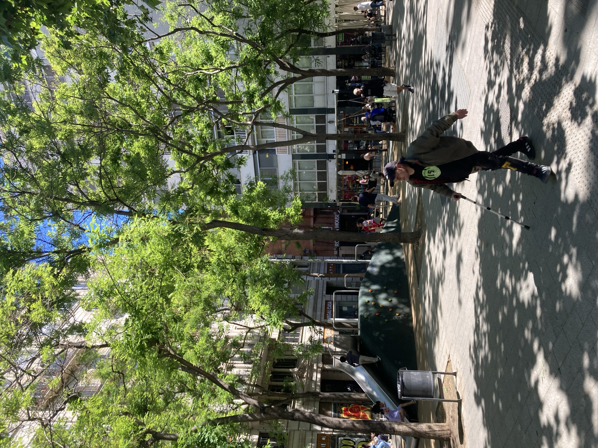 A street scene outdoors in Unknown, Unknown shows a mix of natural and urban elements. The foreground features lush green trees, providing shade and greenery. A playground with a slide is visible. In the background, buildings line the street, with shops and restaurants. People are seen walking and socializing in front of the buildings. A man in a jacket and black pants, wearing a baseball cap and holding a cane is walking on the sidewalk. The sidewalk is paved with patterned square pavers and shaded by tree canopy.