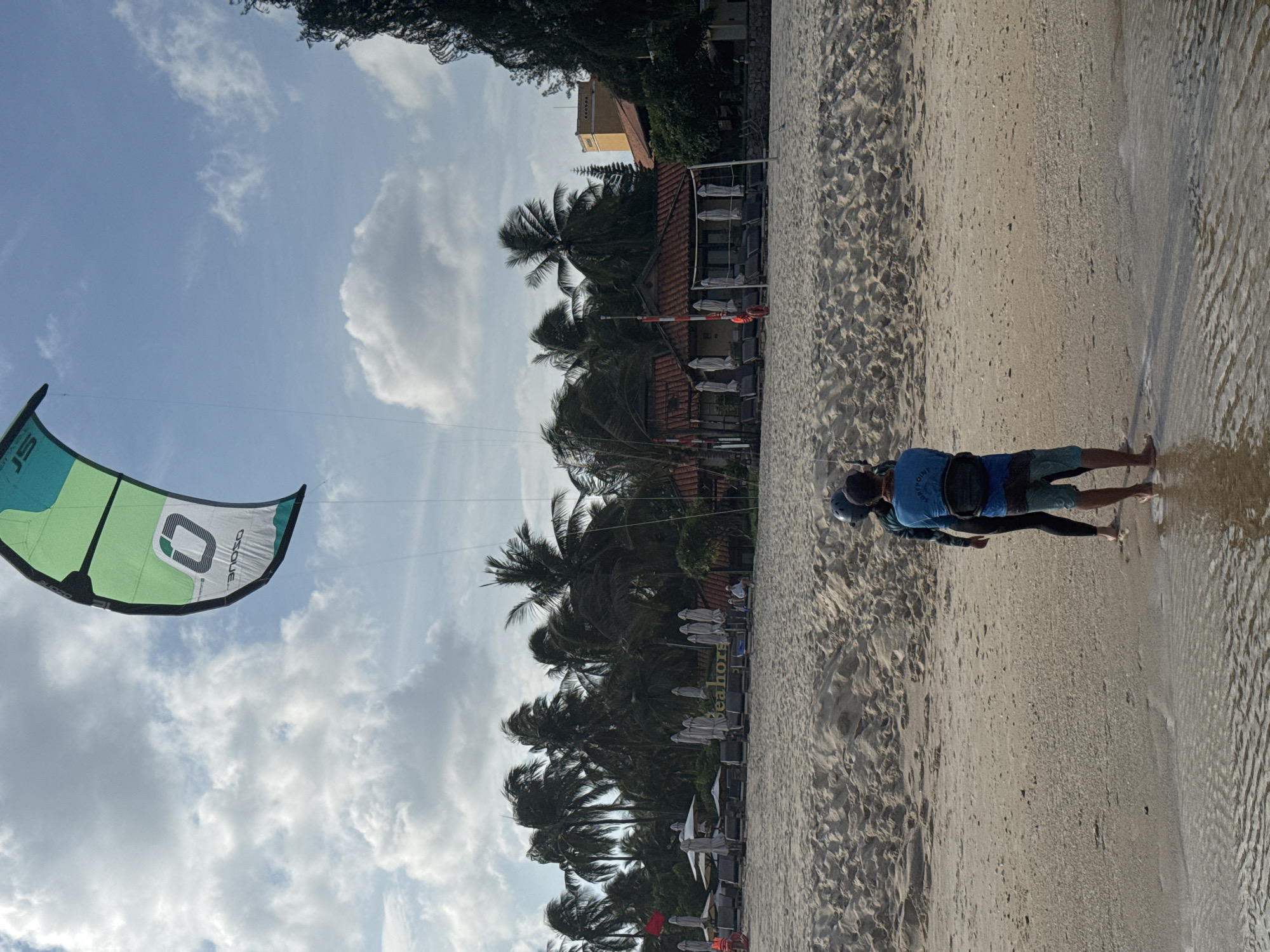 A kitesurfer is preparing to launch on a sandy beach in Mũi Né, Vietnam. The kite, a large green and white object, is inflated and suspended in the air, tethered to the kitesurfer by thin lines. The kitesurfer, wearing a helmet and a blue shirt over a wetsuit, stands on the wet sand at the edge of the water, holding onto the control bar. Another person, possibly an instructor or companion, stands beside the kitesurfer, facing the kite. In the background, palm trees sway in the breeze, and a resort with red-tiled roofs is visible. Beach chairs and umbrellas are set up on the sand, indicating a recreational area. The sky is partially cloudy, with patches of blue sky visible.