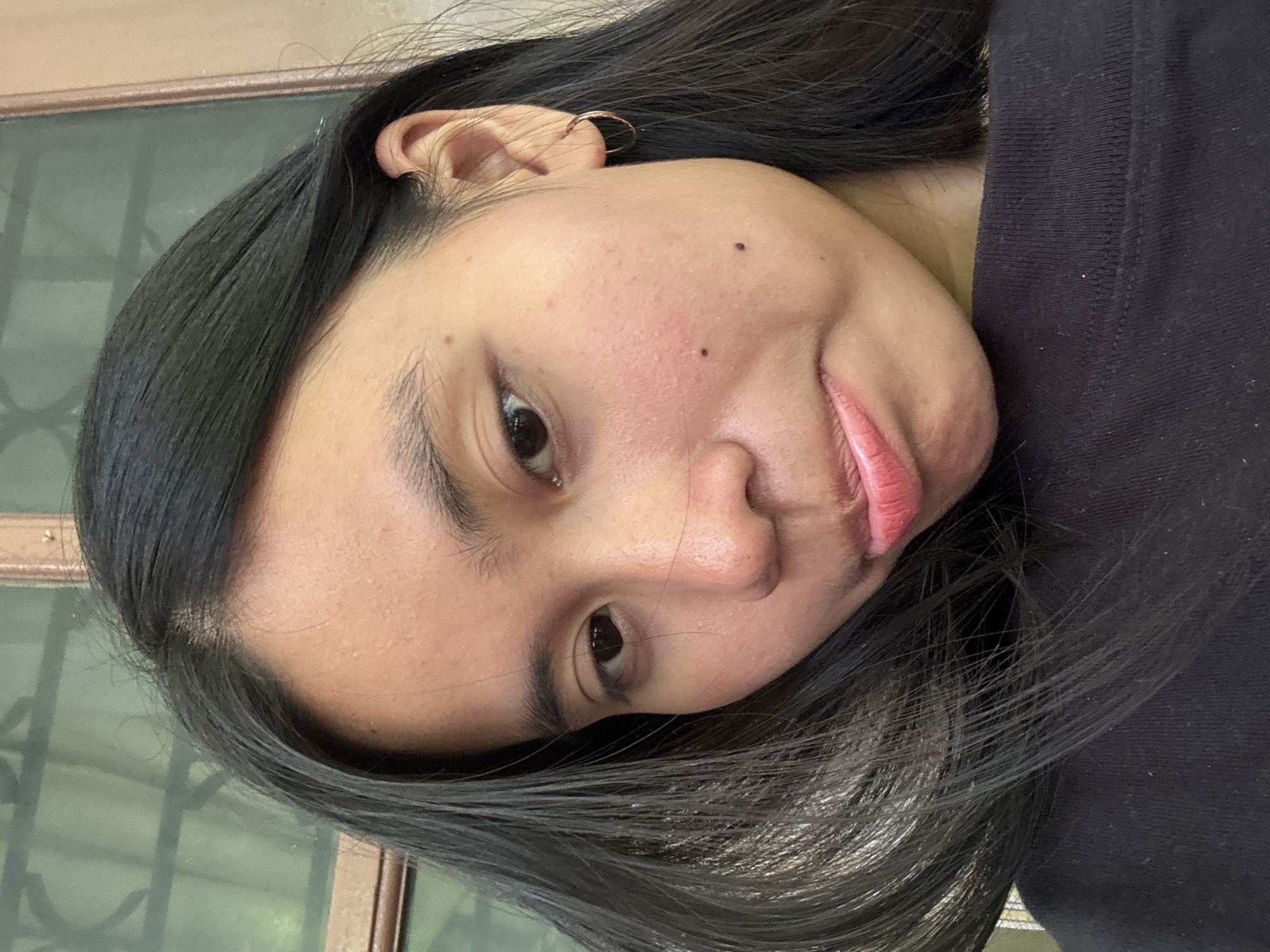A young woman with long, dark hair is shown in a close-up, selfie-style photograph. Her hair is styled to frame her face, with some strands falling over her left eye. She is wearing a black top. Her skin tone is visible, with some light blemishes on her forehead and cheeks. She has a small hoop earring in her right ear. Her lips are slightly pursed and appear to have a subtle pink lipstick applied. The background suggests an outdoor setting, possibly a porch or patio, with a blurred green window or screen and a beige-colored wall or doorframe visible. The overall lighting suggests it is daytime. The image appears to be taken in South Jakarta, Indonesia.