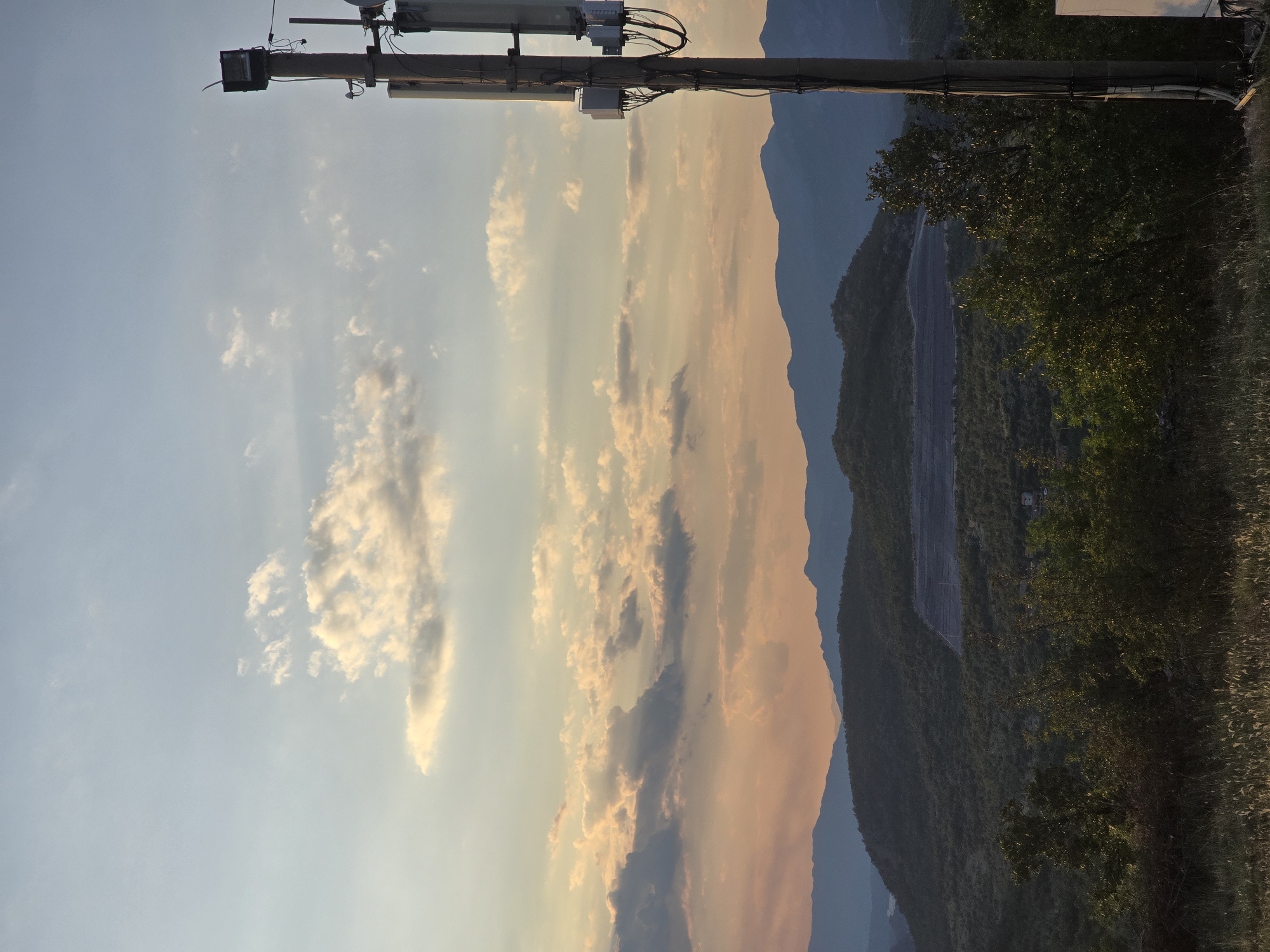 This is a vertical, eye-level, medium shot of a cell tower against a sunset sky over a hilly landscape. The cell tower, dominating the right side of the frame, is made of concrete and metal and is equipped with multiple antennas and a spotlight. The sky, filling the upper half of the image, transitions from a pale blue at the top to a warm orange and pink hue near the horizon, with scattered white and gray clouds. Below the sky, layered mountains fade into the distance, covered in lush green vegetation. In the foreground, a grassy hill slopes down, with some greenery and dry grass. The overall scene depicts a rural or semi-rural setting at dusk, with a man-made structure juxtaposed against a natural landscape.