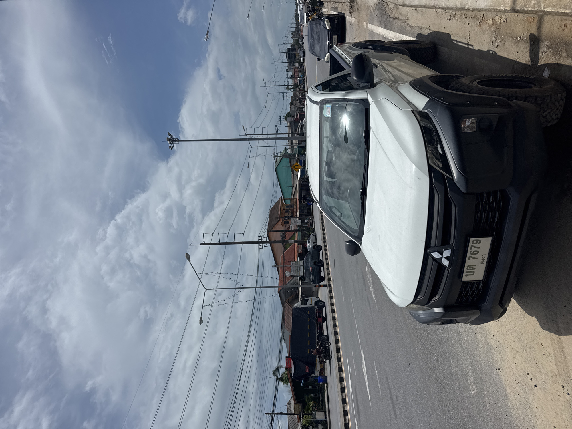 A white Mitsubishi Triton pickup truck is parked on the side of a road in Ban Bang Niang, Thailand. The truck has a black grille and fender flares, and a license plate that reads "บต 7679 พังงา". The road is a multi-lane asphalt road with a sidewalk made of sand and dirt. Overhead power lines and streetlights are visible, as are several buildings along the roadside. In the background, a cloudy sky dominates the scene. Other vehicles, including a black SUV, a white sedan, and a truck with a black tarp, are also present on the road. A person is riding a motorcycle in the distance. The environment appears to be a commercial or mixed-use area in a tropical climate.