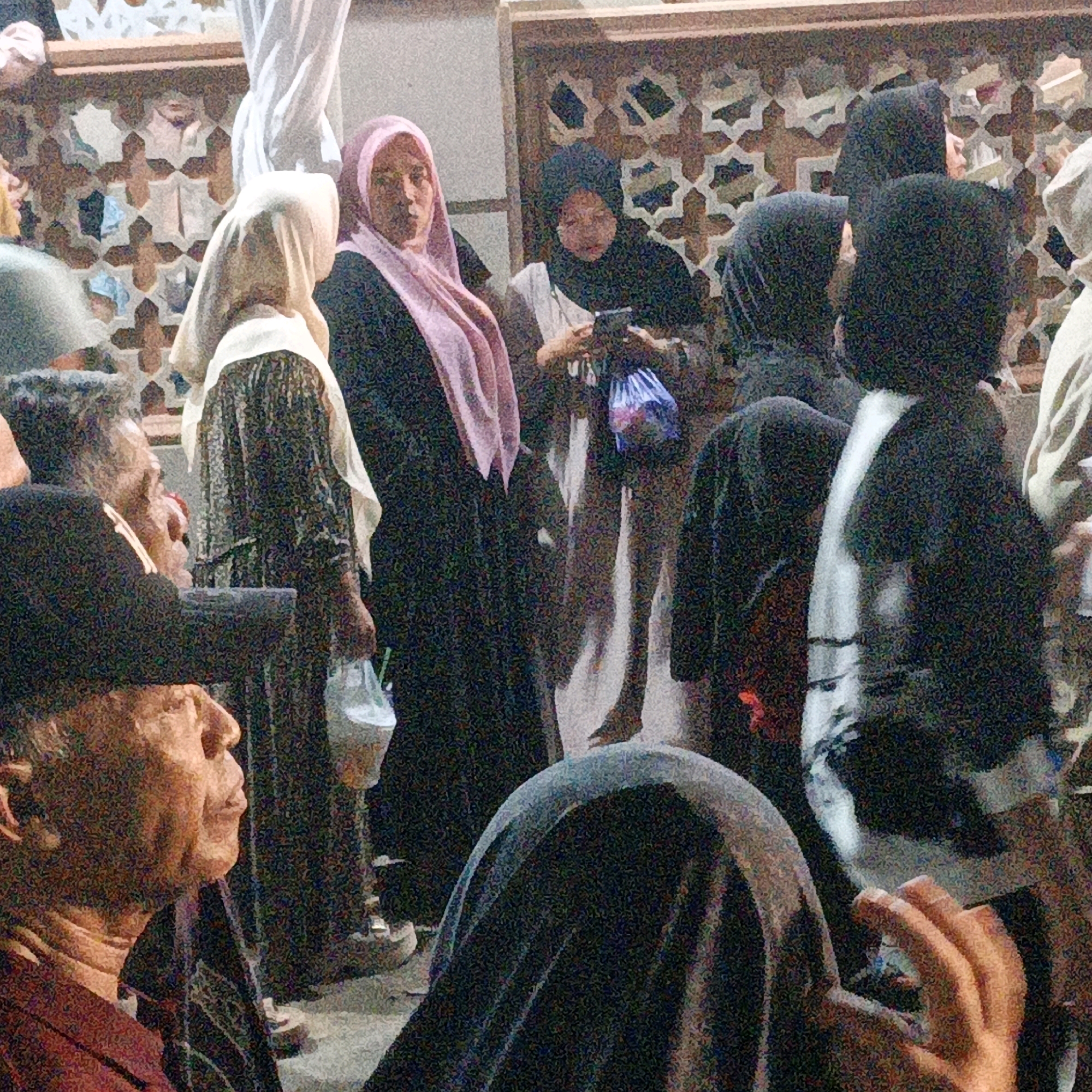 The image depicts a crowd of people, likely at an outdoor gathering, in Kalitorong, Indonesia. The foreground is dominated by the heads and shoulders of several individuals, some wearing traditional Islamic attire such as hijabs. To the left, a man's profile is visible, his facial features appearing weathered and his skin tanned. He is looking to the right, towards other people in the scene. In the center, a woman in a pink hijab is looking forward, her expression difficult to discern due to the image quality. Behind her, another woman in a black hijab is partially visible, holding a phone. The background shows architectural elements, possibly part of a building or a decorative screen, with a geometric pattern. Several people are holding bags, suggesting they might be at a market or event where items are being sold or distributed. The overall lighting suggests it is either early morning or late evening, or possibly an indoor area with artificial lighting. The grainy texture of the image indicates a photograph taken with a lower resolution or in low light conditions.