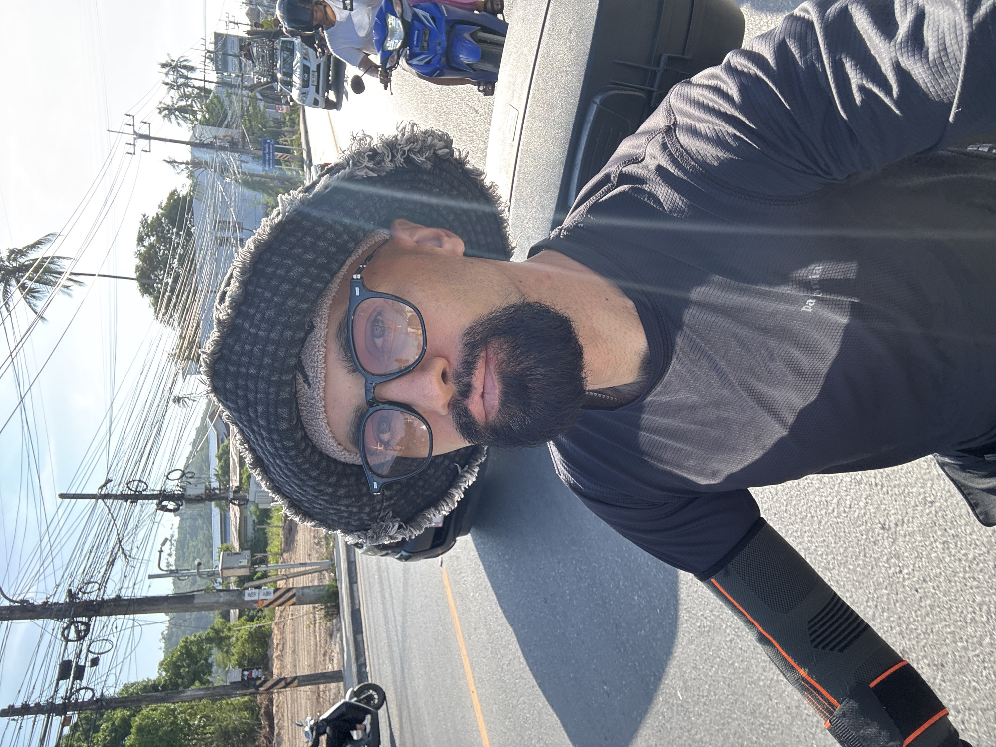 A close-up, selfie-style shot shows a man riding a motorcycle on a sunny day in Ko Samui District, Thailand. The man wears a dark gray bucket hat, sunglasses with orange tinted lenses, and a black long-sleeved shirt. He has a beard and mustache. An orange and black arm sleeve is visible on his left arm. The motorcycle has a black storage box behind the rider. The background depicts a street with utility poles and numerous electrical wires, palm trees, and other motorcycles and vehicles. The road has a yellow line marking.