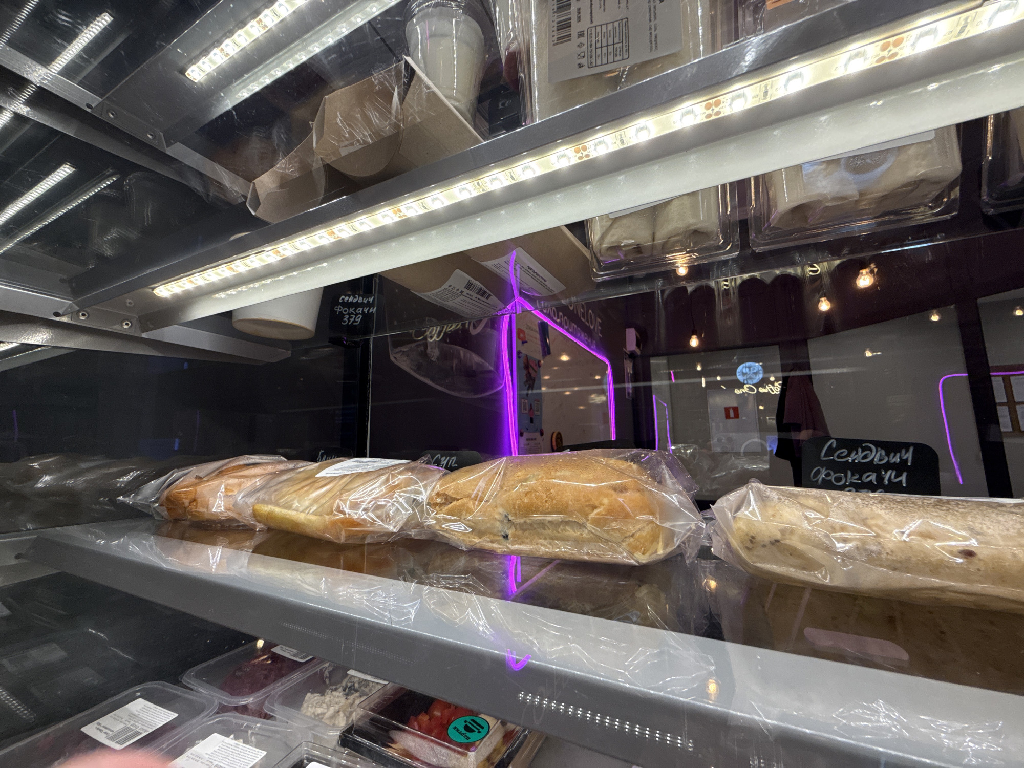 The image depicts a display case of food items, likely in a store or cafe. In the foreground, there is a shelf with packaged sandwiches. Below them are containers with various ingredients, such as what appears to be salads or fillings. The food items are wrapped in clear plastic. Above the sandwiches, there are other food items stacked on shelves under a bright light.
The image is taken indoors with reflections in the glass, which shows a street scene outside. There are lights and signs visible in the reflection, indicating the scene is in an urban area. A sign with the words "Сендвич Фокаччи" (Sandwich Focaccia) is visible. Based on the Cyrillic writing and general style, the location is likely in Moscow, Russia. There are no people visible in the clear view of the image, but reflections suggest some may be outside.