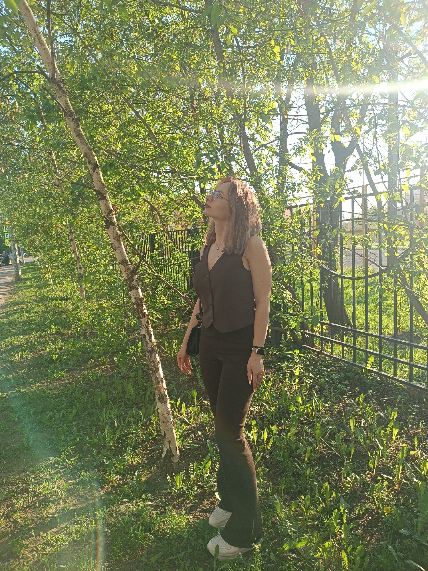 A woman stands in a park-like area in Unknown, Unknown. She is wearing a brown vest and pants, with white sneakers. A small black purse hangs at her side. Her gaze is directed upward toward the trees. Behind her, a black metal fence runs along the edge of the area. Several birch trees line the path, and green foliage fills the space between the path and the fence. The sun is shining, casting shadows and creating a bright, airy feel.