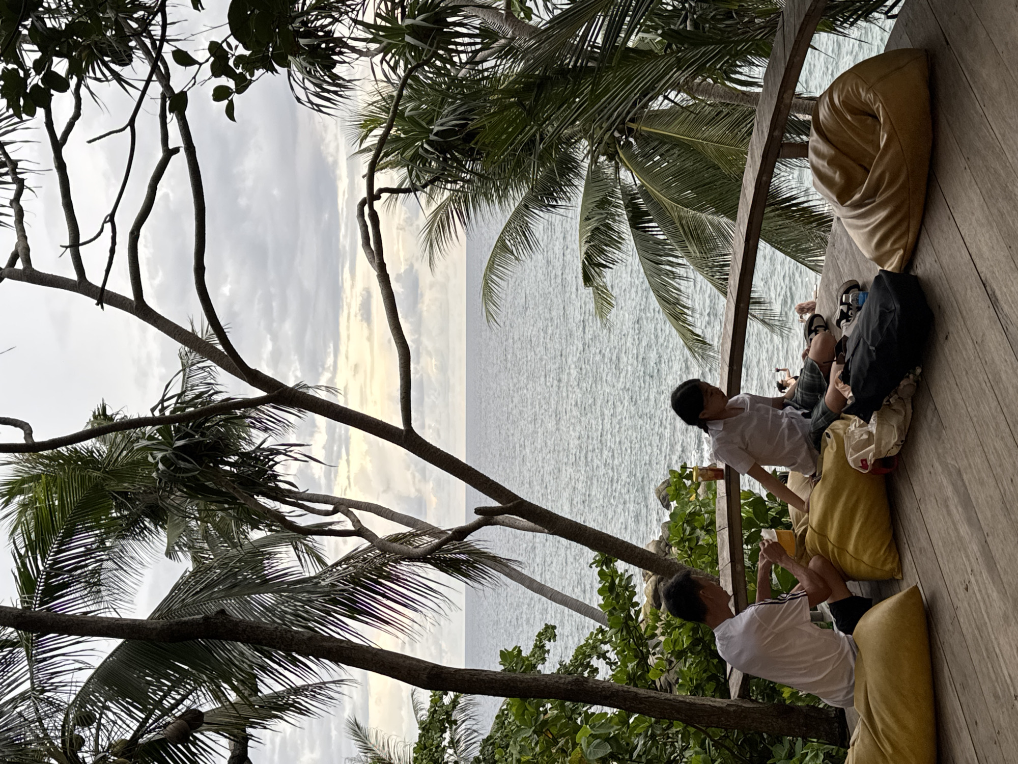 This image depicts a serene outdoor seating area overlooking the ocean, likely in Karon, Thailand. Two individuals, a man and a woman, are seated on large, mustard-yellow beanbag chairs on a wooden deck. The man, wearing a white t-shirt and black shorts, is holding a yellow cup and appears to be interacting with something in his lap. The woman, in a white shirt and plaid pants, is looking down and resting her chin on her hand. Several other beanbag chairs are scattered around the deck, along with bags and shoes. Palm trees and lush foliage frame the scene, with the calm, grey ocean extending to the horizon under a cloudy sky. In the background, two other figures can be seen on the beach, and a yellow drink with a straw sits on the railing. The atmosphere suggests a relaxed and tranquil environment, possibly a cafe or resort lounge.