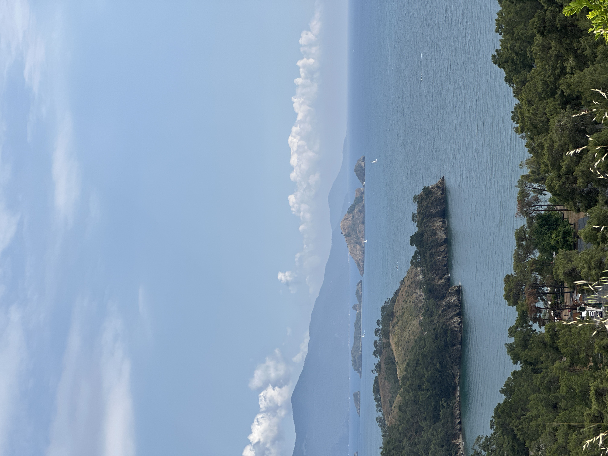 The image depicts a coastal scene in Fethiye, Turkey, featuring a series of small, green islands extending into the water. The sea transitions from a lighter shade near the horizon to a deeper blue closer to the viewer. White sailboats are scattered on the water. Mountains rise from the sea, covered with vegetation. Fluffy white clouds are present in the blue sky. The scene is bordered by dense trees on the right edge.