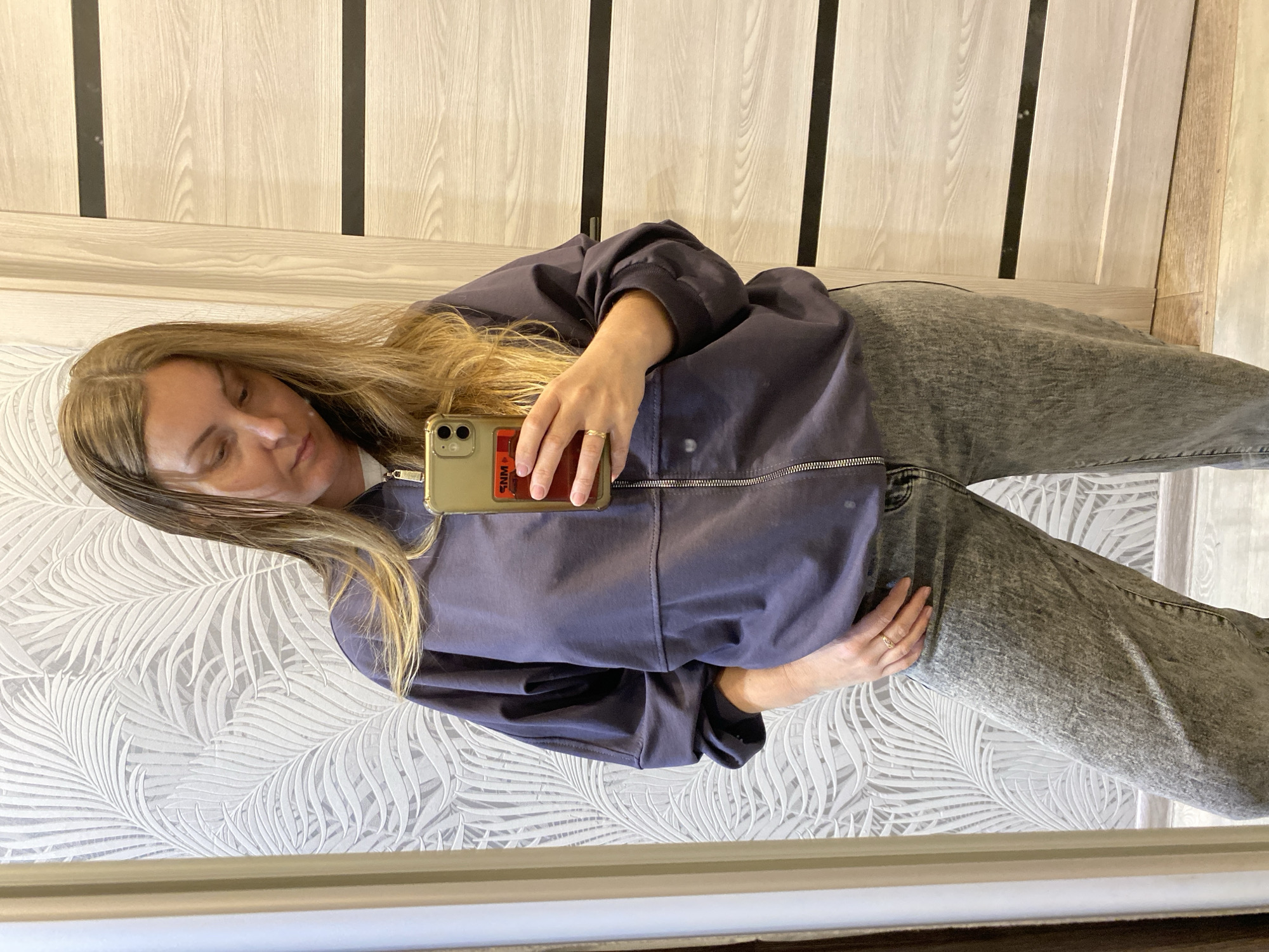 A person is taking a selfie in a mirror. The person is wearing a dark purple bomber jacket and grey jeans. The person is holding a gold iPhone. The background includes a wall with a leaf pattern wallpaper and a door or wall panel with vertical stripes. There is no evidence to suggest that the location is in Noviy, Russia.