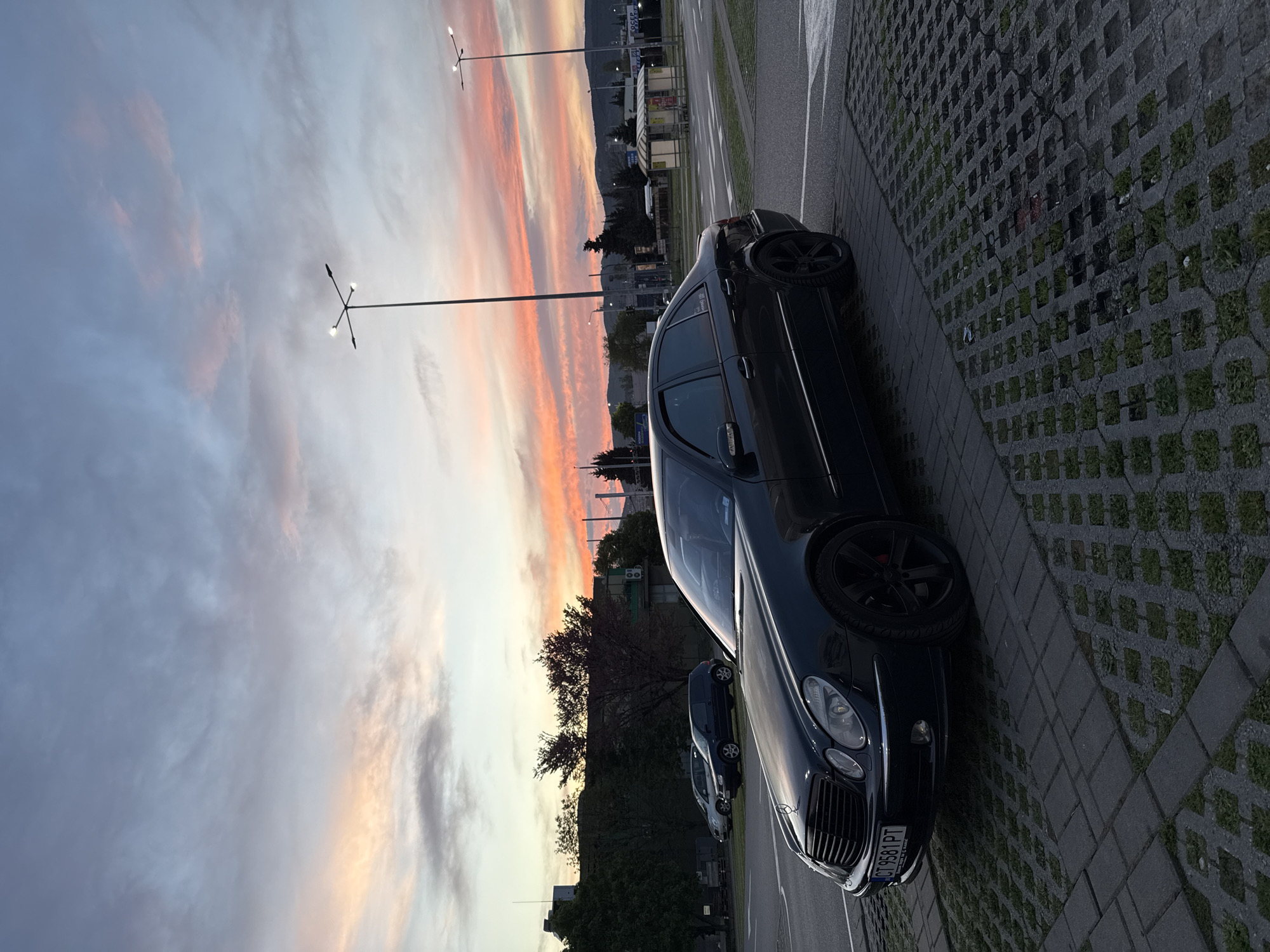 The image depicts a black Mercedes-Benz E-Class parked on a patterned concrete parking lot. The location appears to be in Stara Zagora, Bulgaria, based on the license plate visible on the vehicle. The car is positioned close to a curb adjacent to a street. A sunset sky with orange and pink hues is visible in the background, along with distant structures. The scene is illuminated by natural light. Other parked vehicles are visible in the background. Streetlights are also visible.