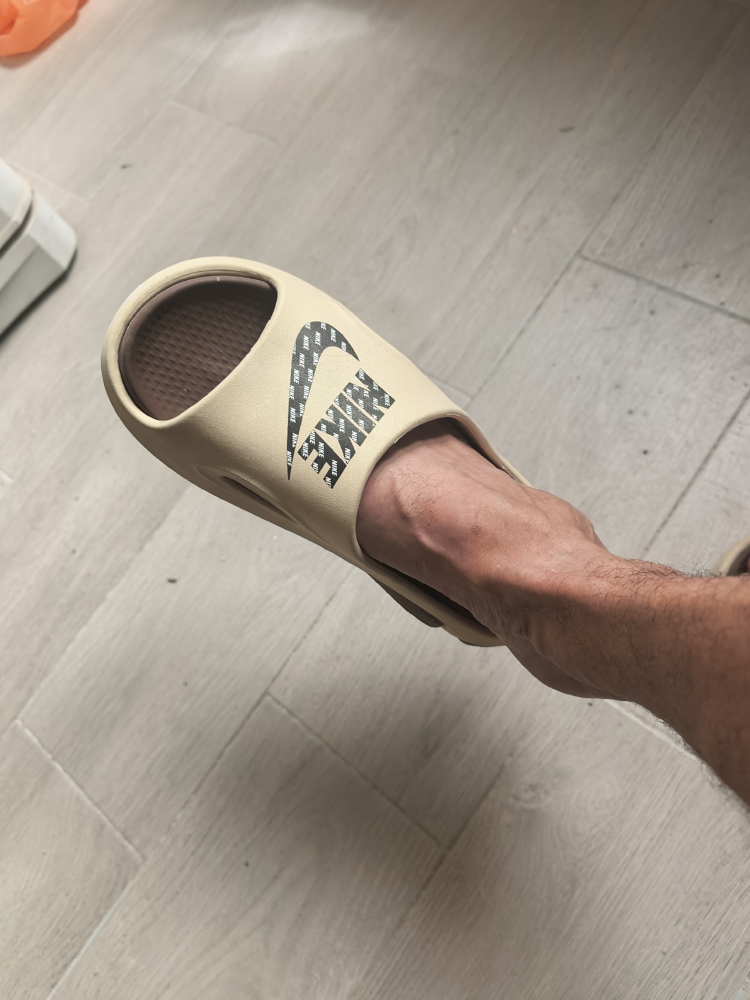 The image shows a close-up view of a person's foot wearing a tan Nike slide on a light-colored, wood-plank floor. The slide has a Nike logo on the strap, which is made up of repeated "NIKE" text in black. The foot is bare and has visible veins and hair on the ankle and lower leg. The background is out of focus, but appears to be an indoor setting with a wall to the left and a floor with visible grout lines between the planks. There are some small dark specks on the floor, possibly dust or dirt. The lighting suggests an indoor environment, possibly during the daytime. There is no specific indication of the time of day or weather conditions. The overall scene is casual and domestic, with the focus on the footwear. Visible text includes the word "NIKE" repeated multiple times within the logo on the slide.