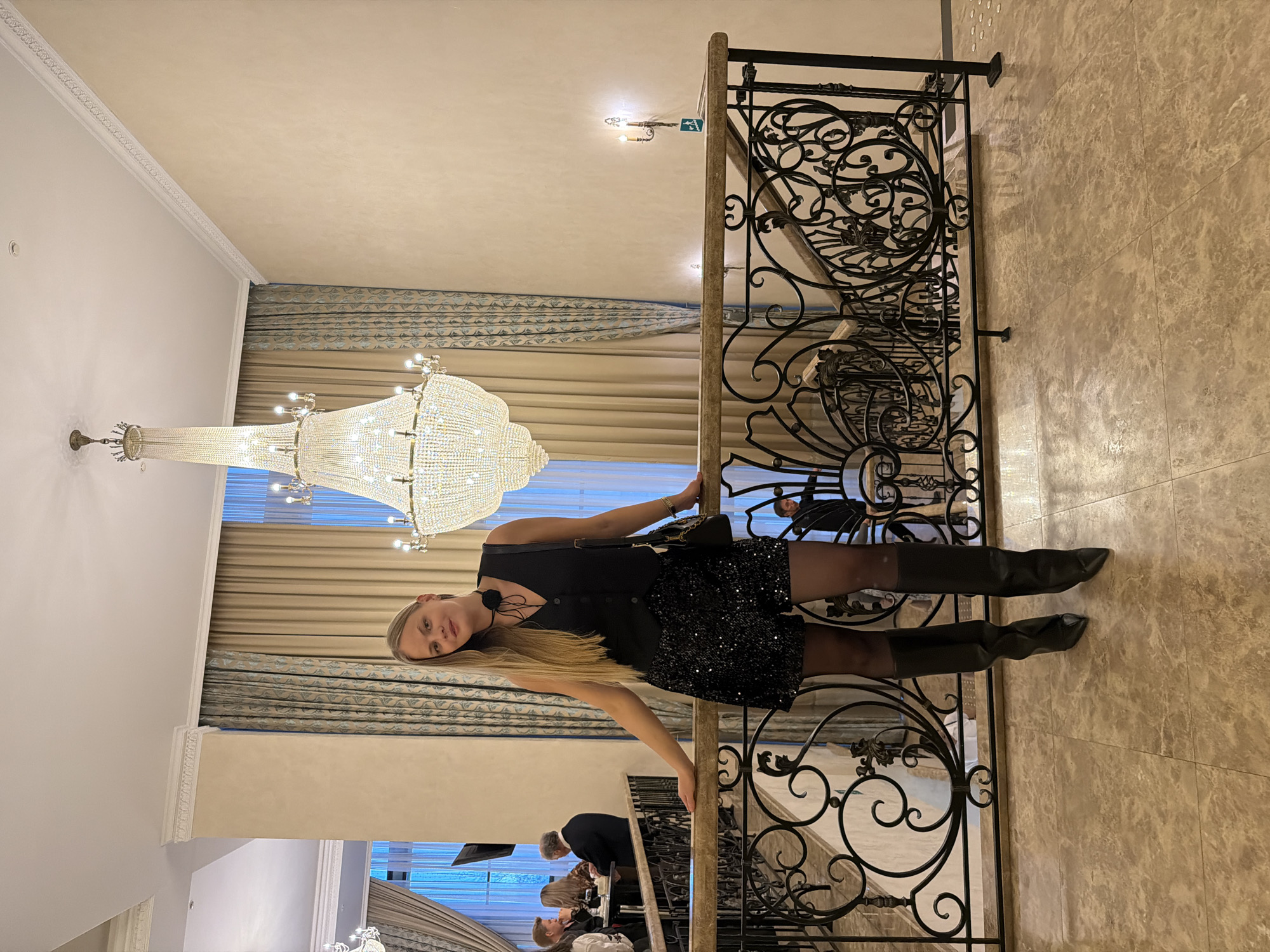 A woman in a black dress and boots stands on a staircase in a building in Izhevsk, Russia. The staircase has an ornate metal railing. Behind her, there is a large chandelier and patterned curtains. Other people can be seen on the lower levels of the staircase. The floor and walls have a tiled finish.