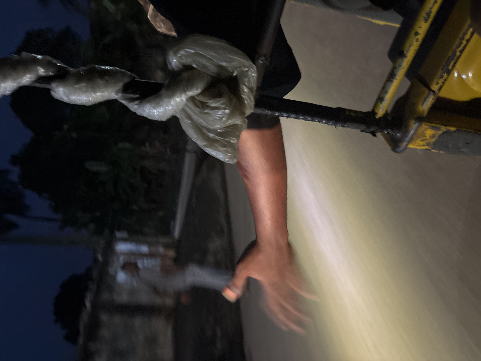 An over-the-shoulder, night shot captures a person's arm and hand reaching out from the right side of the frame. The arm is tanned, with skin appearing flushed from the camera's light source. The hand is open, palm facing slightly upwards, with fingers spread. In the background, a blurry figure in a light-colored garment walks away from the camera on a street. Adjacent to the figure is a low-lying building with a dark roof. The scene appears to be a street in Onitsha, Nigeria, given the humid, tropical vegetation visible as dark silhouettes against the night sky. A yellow metal frame, possibly part of a vehicle or structure, occupies the lower right corner of the image, with a plastic-wrapped object hanging from a black pole above it. The ground is illuminated by a strong, warm light, suggesting a vehicle's headlights are the primary light source.