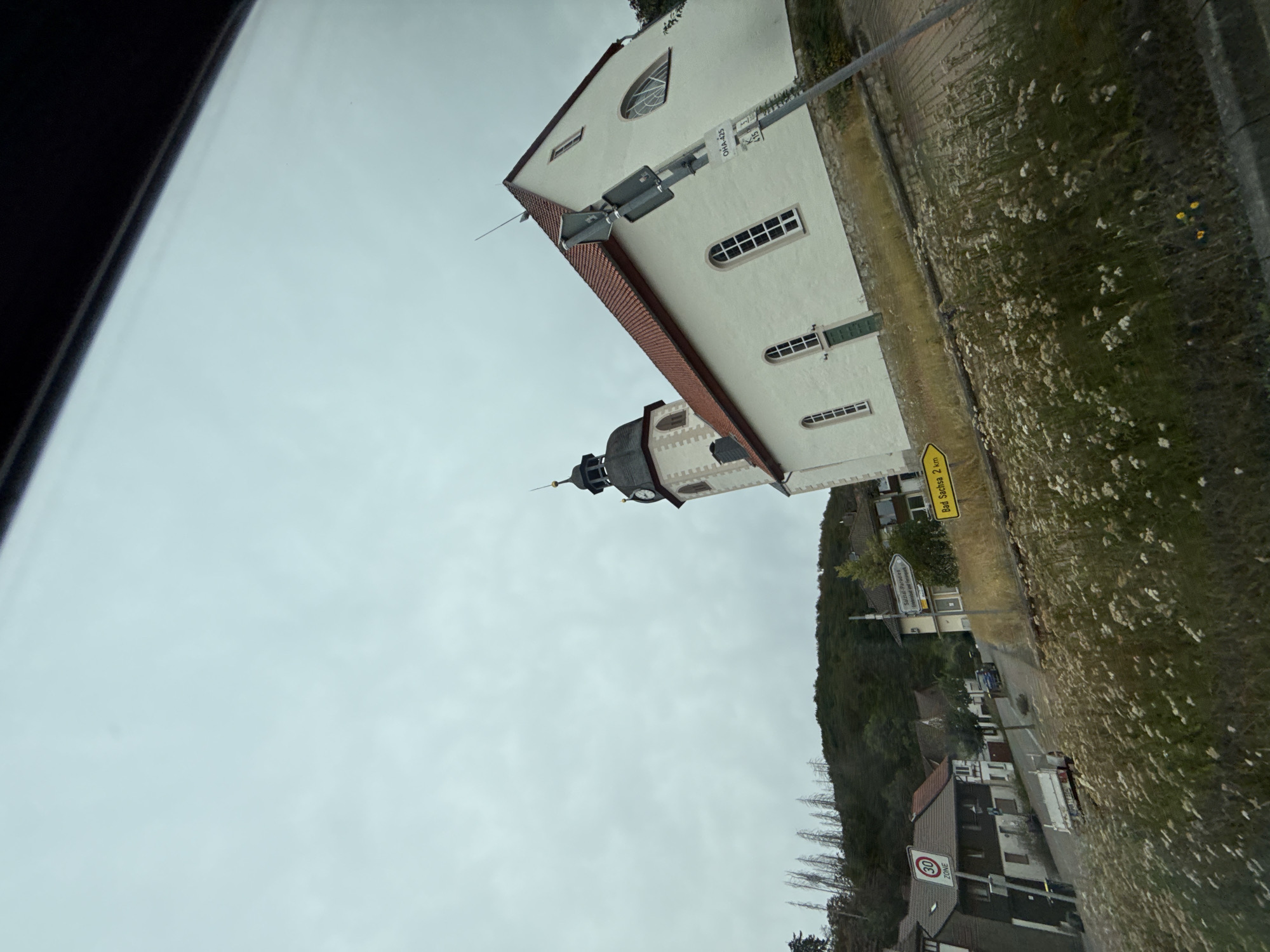 The image shows a view of a white church with a dark steeple and a red roof, located in Bad Sachsa, Germany. The church is situated on a hill, with a densely wooded hillside behind it. In the foreground, there is a street with a few houses and a signpost indicating "Bad Sachsa 2 km". The sky is overcast with a light grey cloud cover. There are no people visible in the image. The overall scene suggests a rural or semi-rural setting with a focus on the church as a prominent landmark.