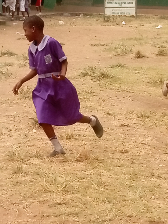 The image displays an outdoor scene at the Community High School Offe in Atani, Nigeria. A young individual, likely a girl, with dark skin and short dark hair, is captured in motion, running or skipping across a dry, sparse field. She wears a purple short-sleeved school dress featuring a white collar, white cuffs, and a white sash at the waist, with a light-colored emblem on the left chest. She wears white socks and dark-colored, closed-toe shoes. Her left foot is elevated, and her right foot is on the ground, indicating movement. Her arms are bent at the elbows, with hands slightly open. She looks to her left with a visible smile. The ground is dry, reddish-brown earth with scattered patches of yellowish-brown grass and weeds. In the upper left background, blurred figures of other individuals in light-colored clothing are partially visible. To the right, a green building or wall is present, alongside a white rectangular sign. Legible text on the sign includes "COMMUNITY HIGH SCH. OFFE," "ATANI," and "HON. AFAN VICTOR UGFNE," confirming the location and context.